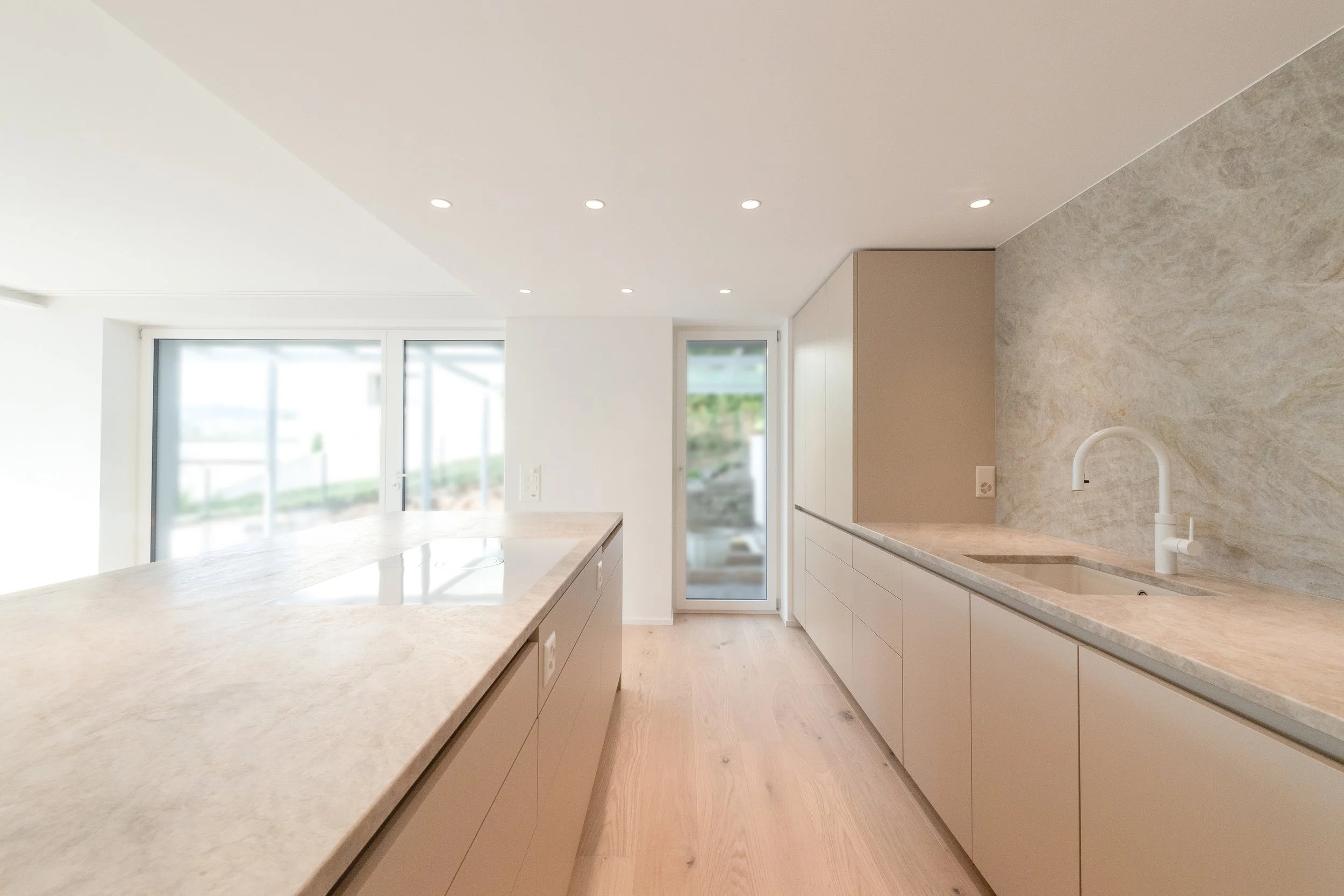 Moderne, helle Küche mit weißen Schränken, Marmorarbeitsplatten, einem Spülbecken mit weißem Wasserhahn, Holzfußboden und großen Fenstern