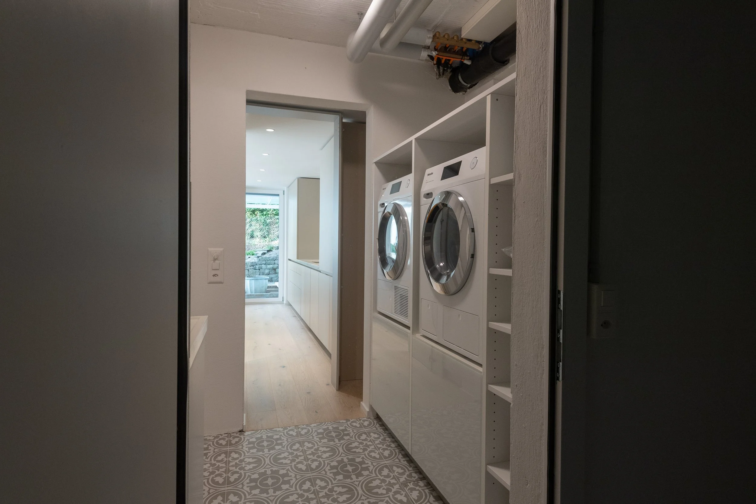 Blick in einen Hauswirtschaftsraum mit Waschmaschine und Trockner, Badezimmerfliesen und einem Übergang zu einem Flur mit Holzfußboden und Blick nach draußen.