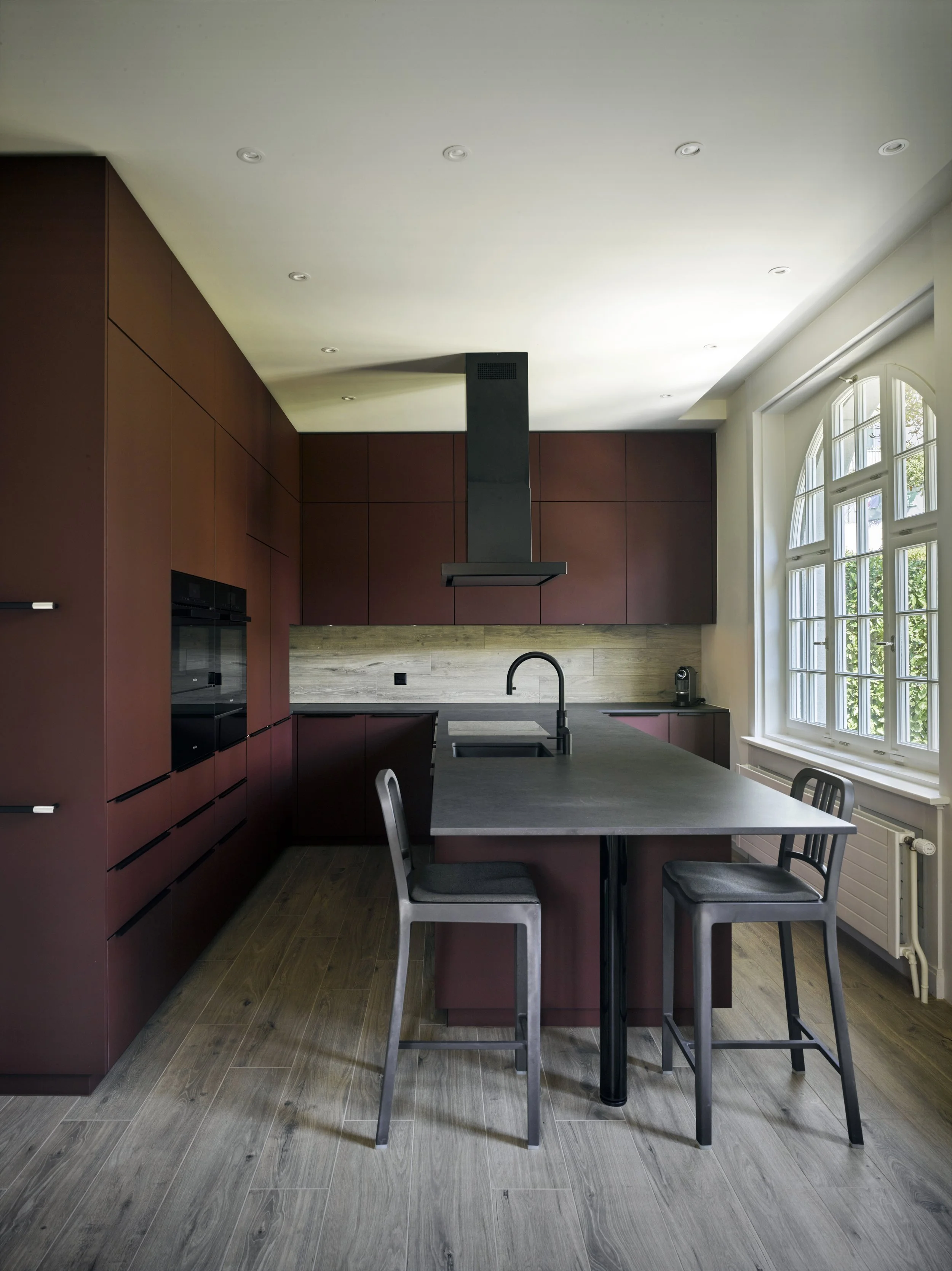 Moderne Küche mit roten Schränken, schwarzer Arbeitsplatte, Fenster mit Bogen, Kücheninsel mit zwei Stühlen, Küchenventilator an der Decke. 
