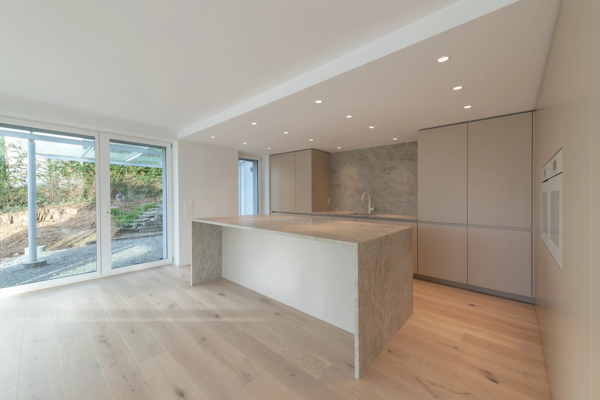 Moderner Küchenraum mit hellen Wänden, einer Kücheninsel aus hellem Stein und einem Wasserhahn. Große Glastüren führen nach draußen, und die Decke ist mit kleinen Einbauspots beleuchtet.
