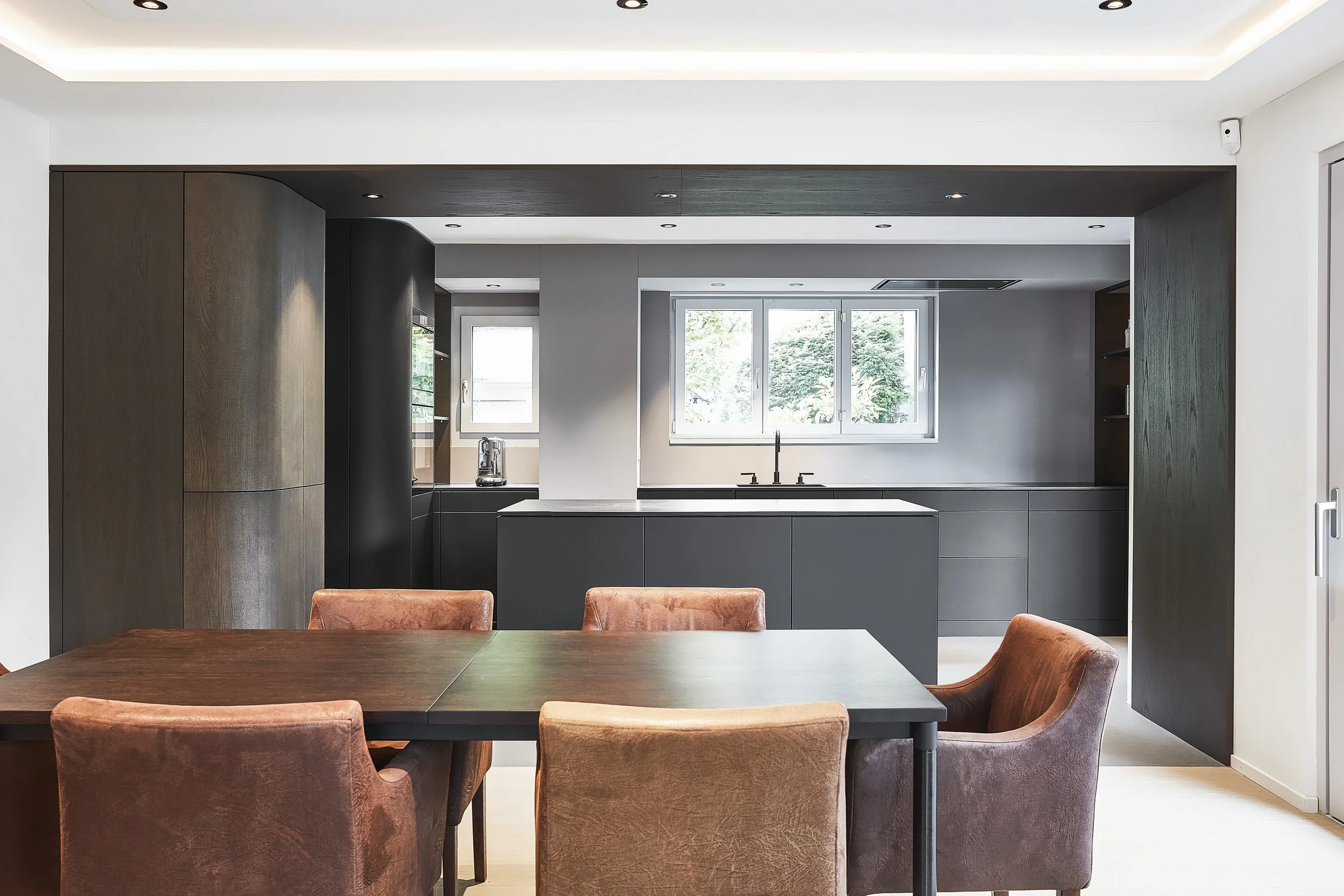 Moderne Küche mit schwarzen Schränken, einem großen Fenster und einem Holztisch mit vier braunen Samtsitzen.