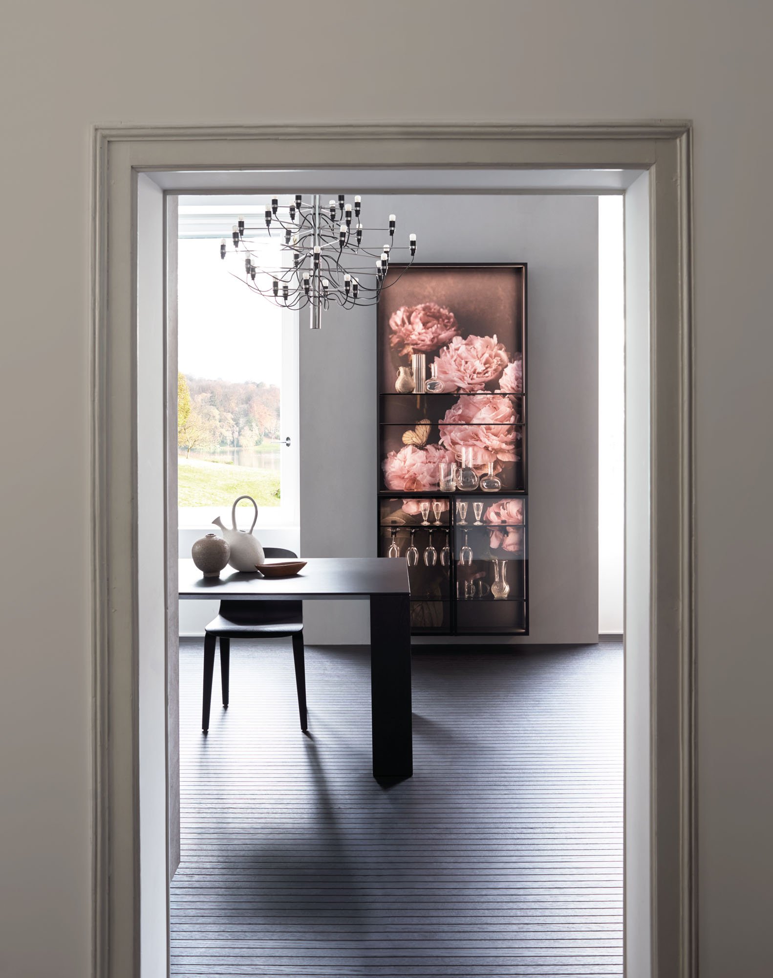 Ein Blick durch eine offene Tür in einen modern eingerichteten Raum mit einem schwarzen Esstisch, Stuhl und einem Regal mit großen pinken Blumenbildern. Es ist Tageslicht und eine Landschaft im Hintergrund sichtbar.