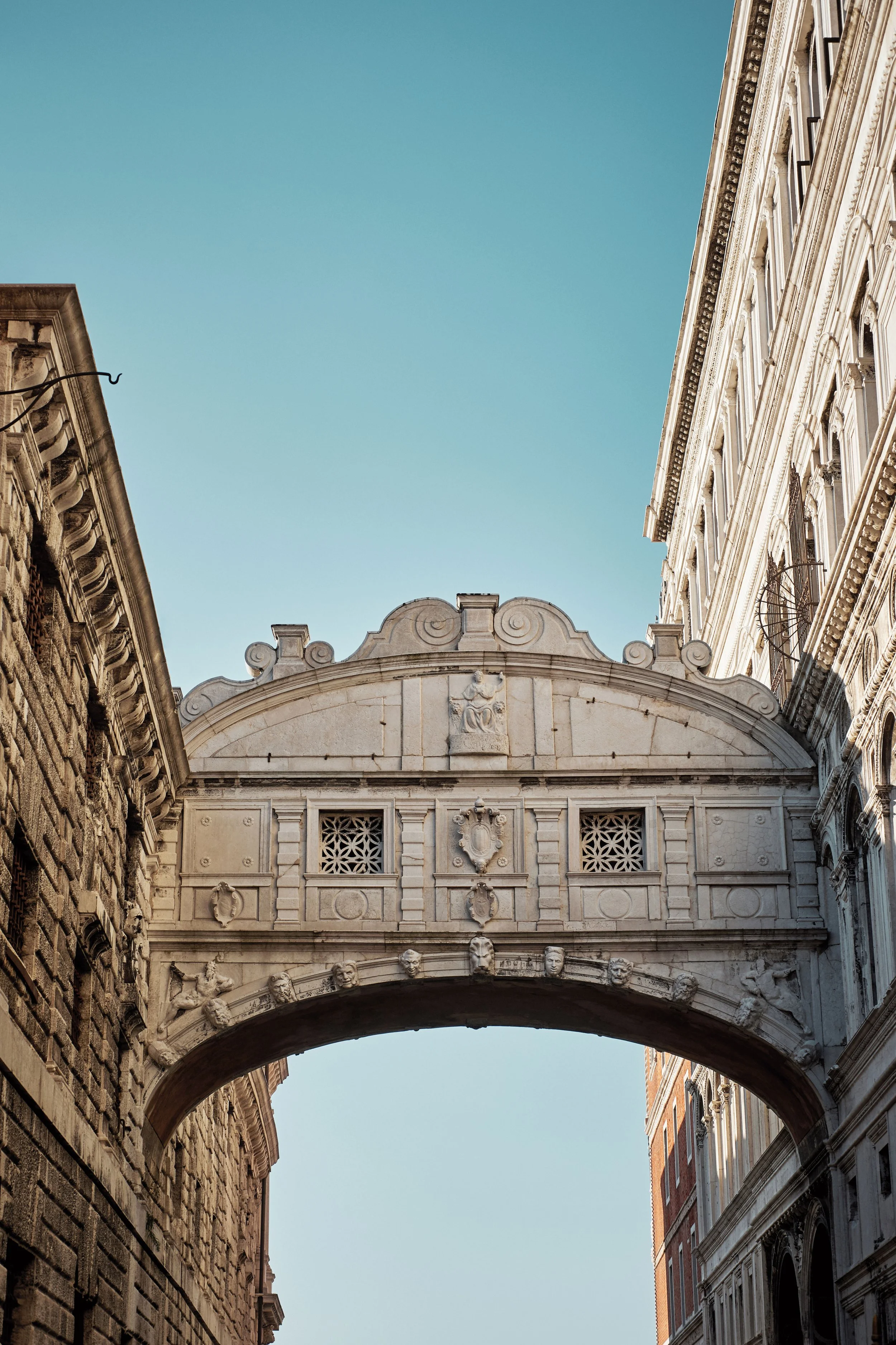 Architektonischer Bogen mit Verzierungen und Gesichtern, umgeben von historischen Gebäuden mit aufwändiger Fassadengestaltung unter blauem Himmel.