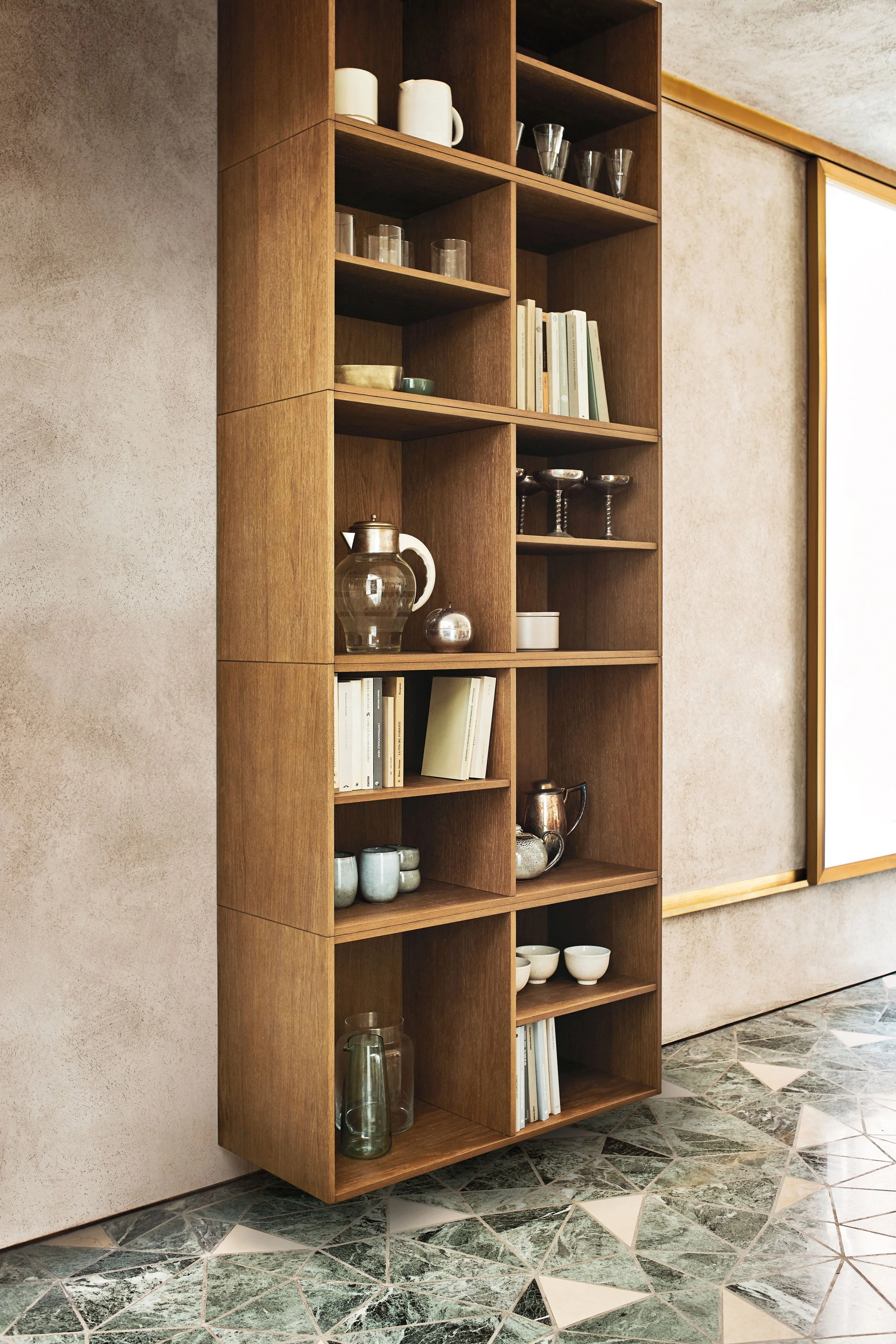 Hölzerner Bücherregal mit Geschirr, Büchern und Dekorationsgegenständen, an einer Wand. Am Boden große Fliesen in Muster aus neutralen Tönen.