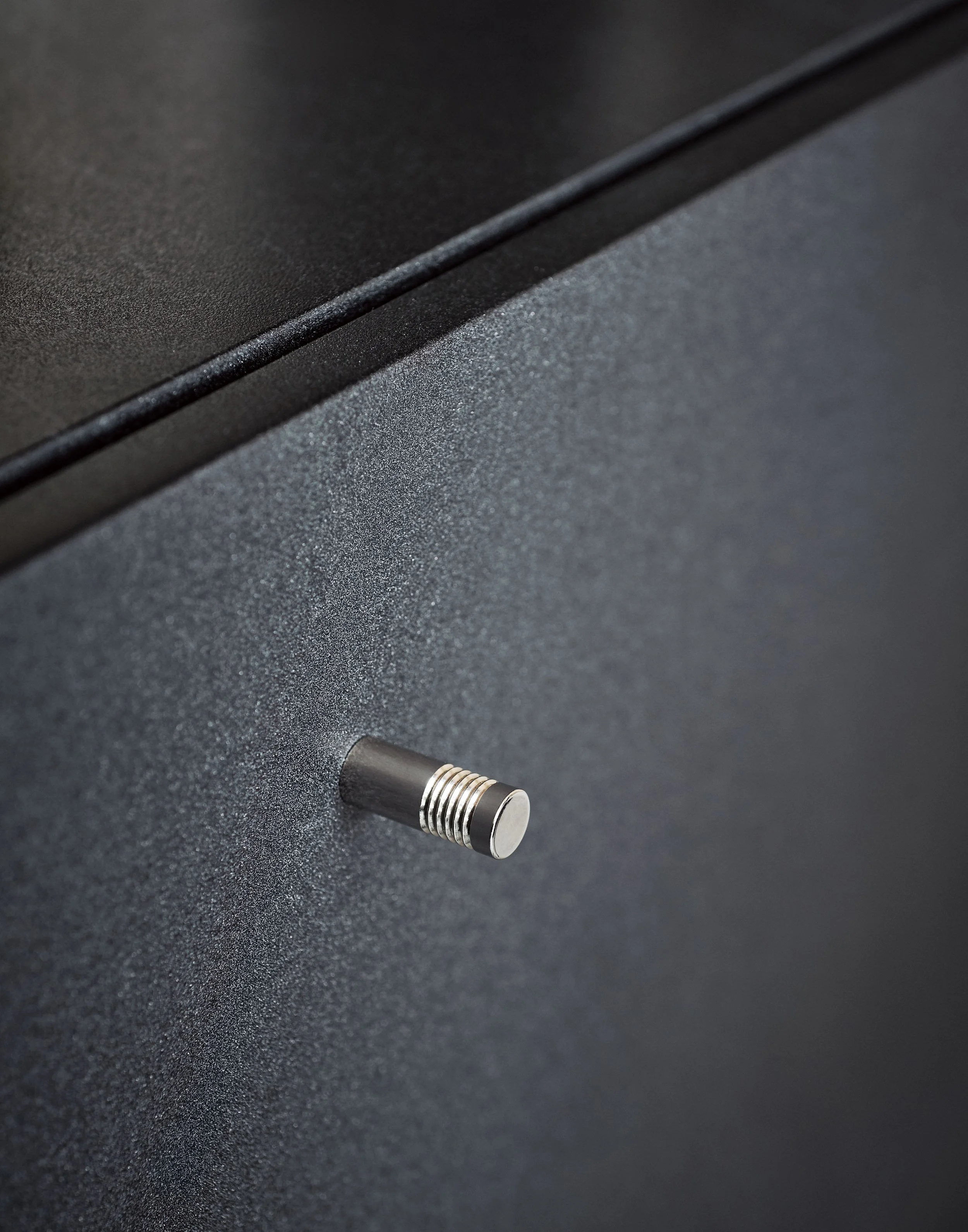 Nahaufnahme eines schwarzen, strukturierten Oberflächen mit metallic-glänzendem Druckknopf im unteren Bereich.