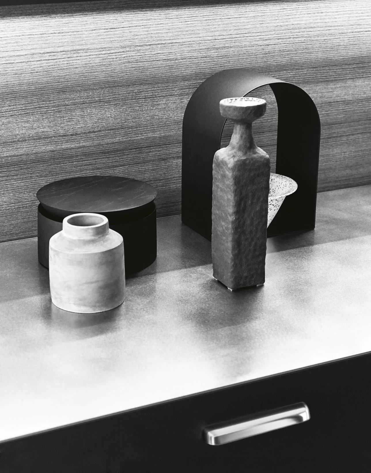 Schwarz-weiß Fotografie eines minimalistischen Stilllebens mit drei dekorativen Objekten auf einem Tisch: einer kleinen hellen Vase, einer organisch geformten Skulptur und einer runden schwarzen Box mit Deckel. Der Hintergrund aus Holz verleiht der S