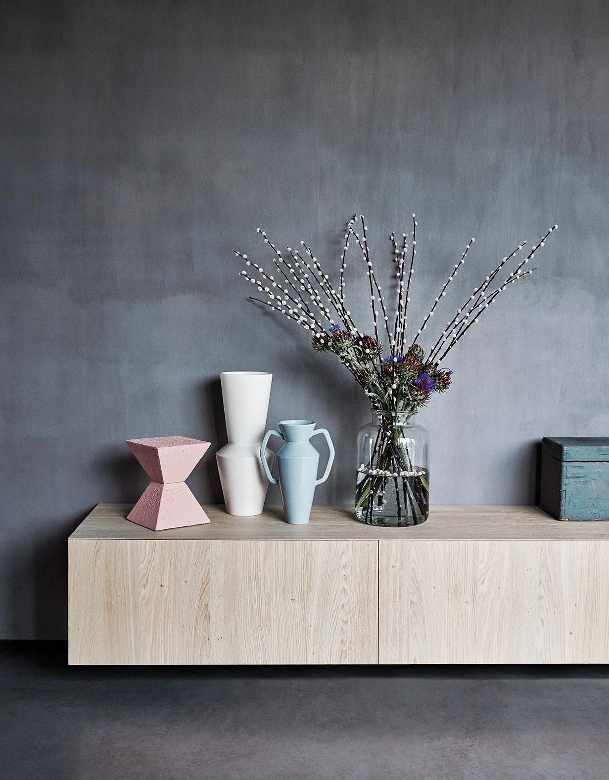 Eine Holzkonsole mit vier Vasen und einer kleinen Kiste. Die Vasen enthalten Absinth, eine graue Vase, eine hellblaue Vase mit Griffen und einen großen Blumenstrauß aus weißen Zweigen und bunten Blumen in einer Glaskaraffe.