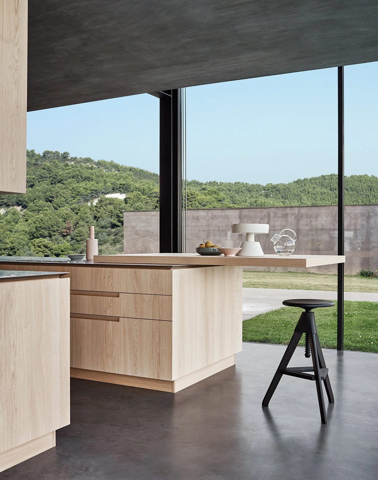 Moderne Küche mit Holzarbeitsplatte, große Fenster mit Blick auf grüne Hügellandschaft, minimalistische Dekoration, schwarzer Hocker im Vordergrund.