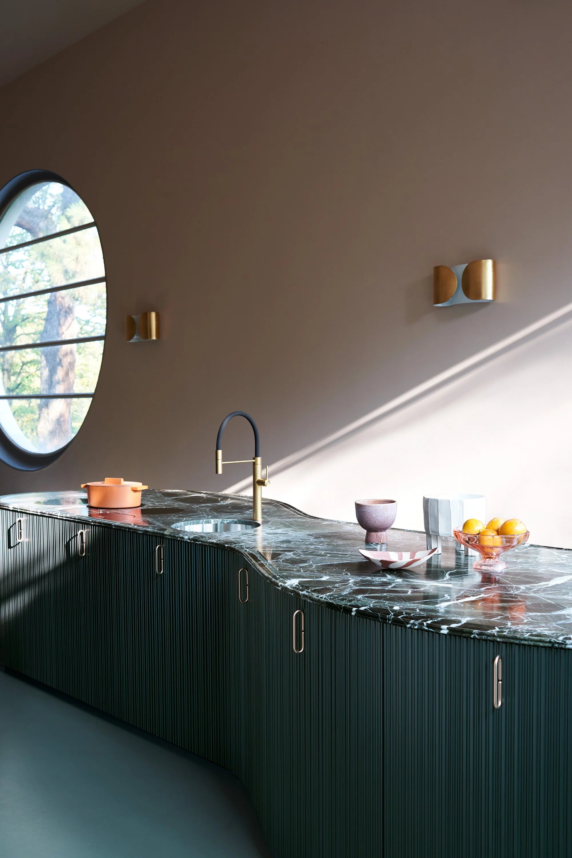 Ein moderner Küchenbereich mit einer schwarz-grünen Marmorarbeitsplatte, goldenen Wasserhähnen, bunten Geschirr und einer großen runden Fensterscheibe, die viel Tageslicht hereinlässt.
