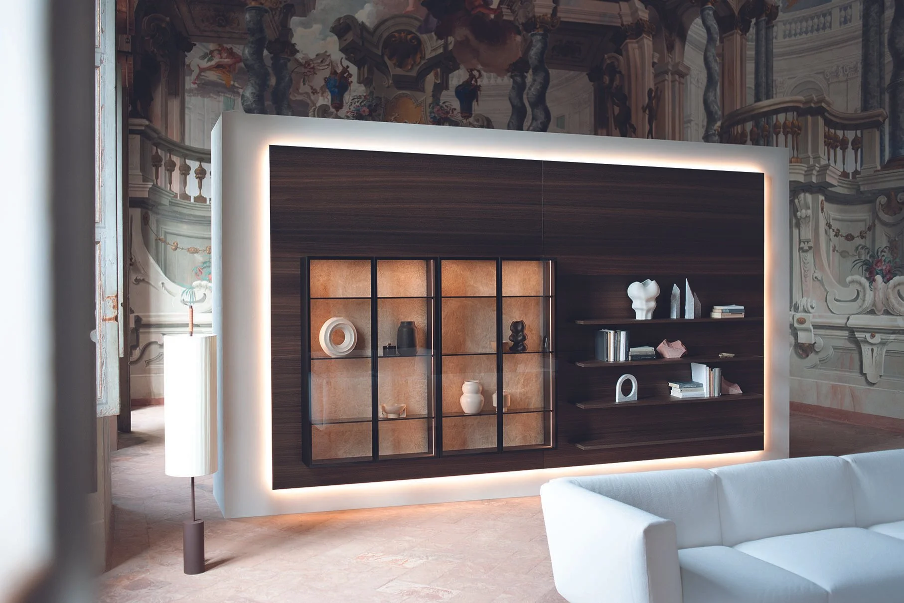 Moderne Holztrennwand mit integrierten Regalen und Beleuchtung, getrennt durch einen weißen Rahmen, im Hintergrund eine alte Wandmalerei im barocken Stil.