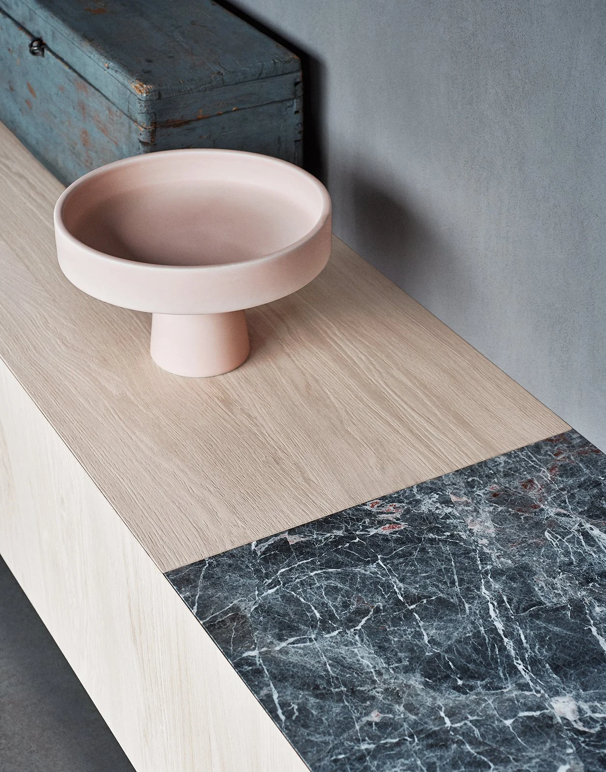 Eine elegante rosa keramische Schale auf einem Holztablett, im Hintergrund ein altmodischer blauer Metallkoffer.