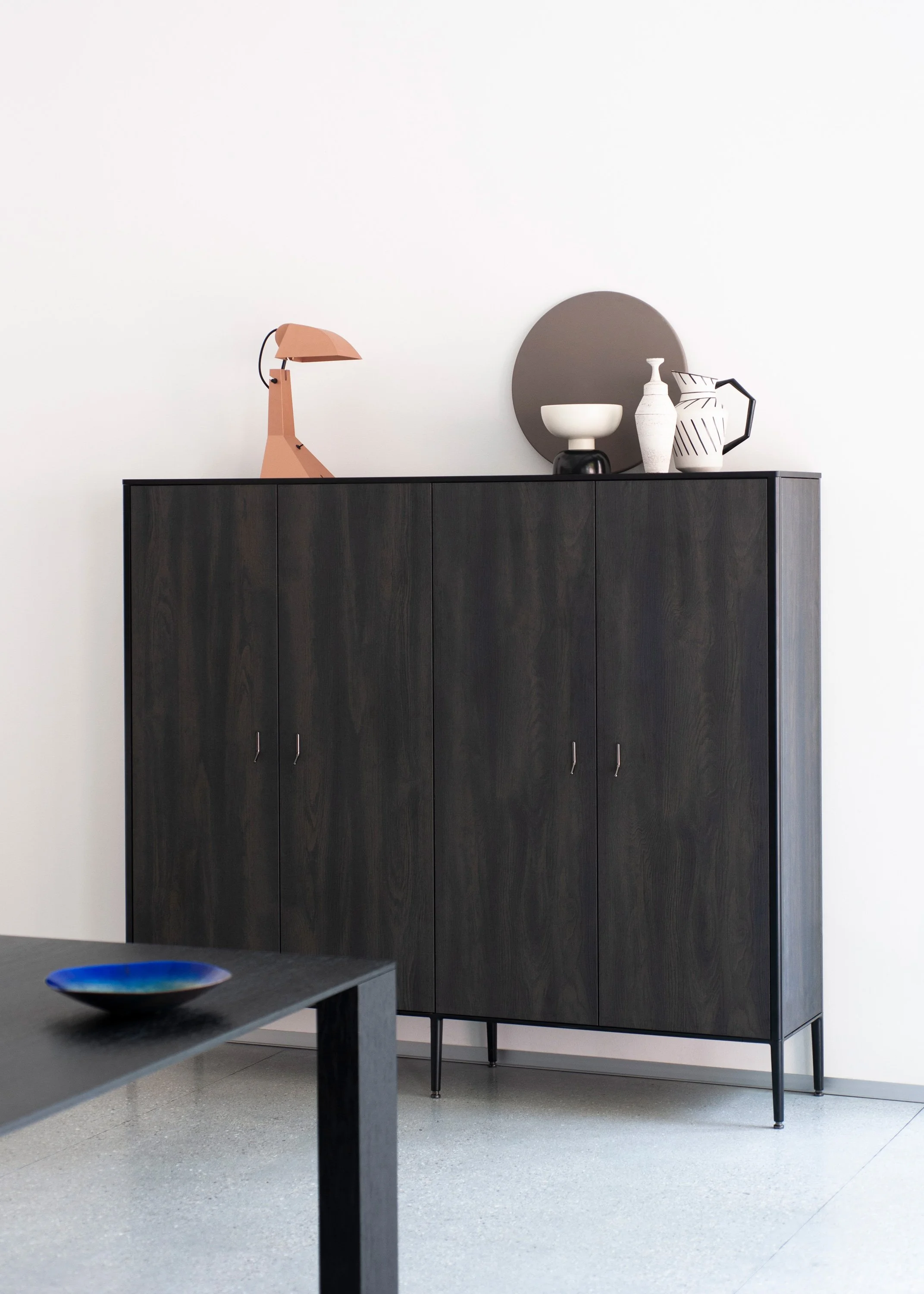 Ein schwarzer Schrank mit Dekorationsgegenständen wie einer weißen Vase, einer schwarzen Vase, einer durchscheinenden weißen Karaffe und einer weißen Vase mit Mustern, vor einem weißen Wand. Im Vordergrund ein grauer Tisch mit einem blauen Tellerrand