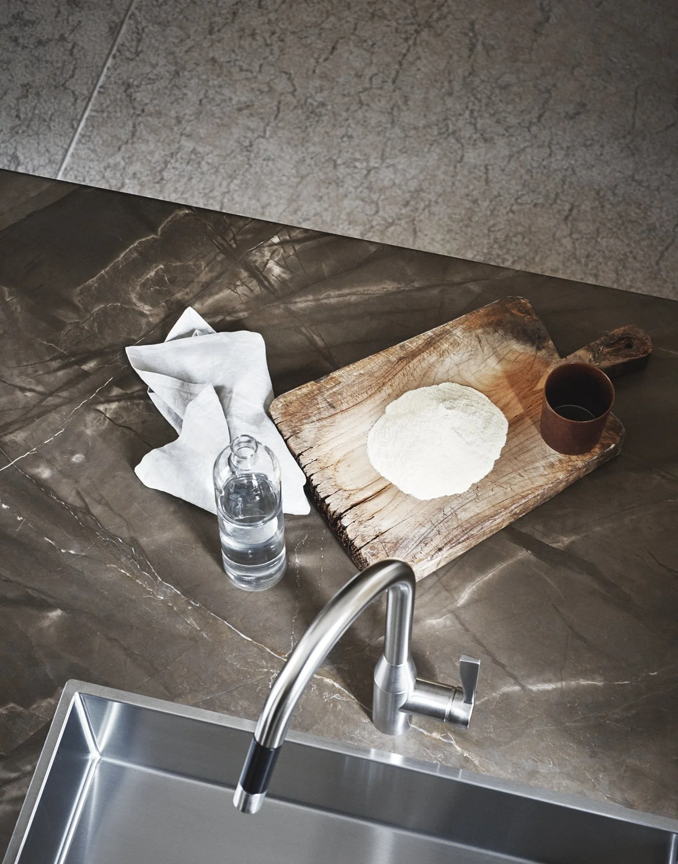 Ein Küchenhintergrund mit einem Wasserhahn, einem Spülbecken, einem Holzbrett mit Mehl und einer Tasse auf einer dunklen Arbeitsplatte, sowie einer weißen Papierserviette.