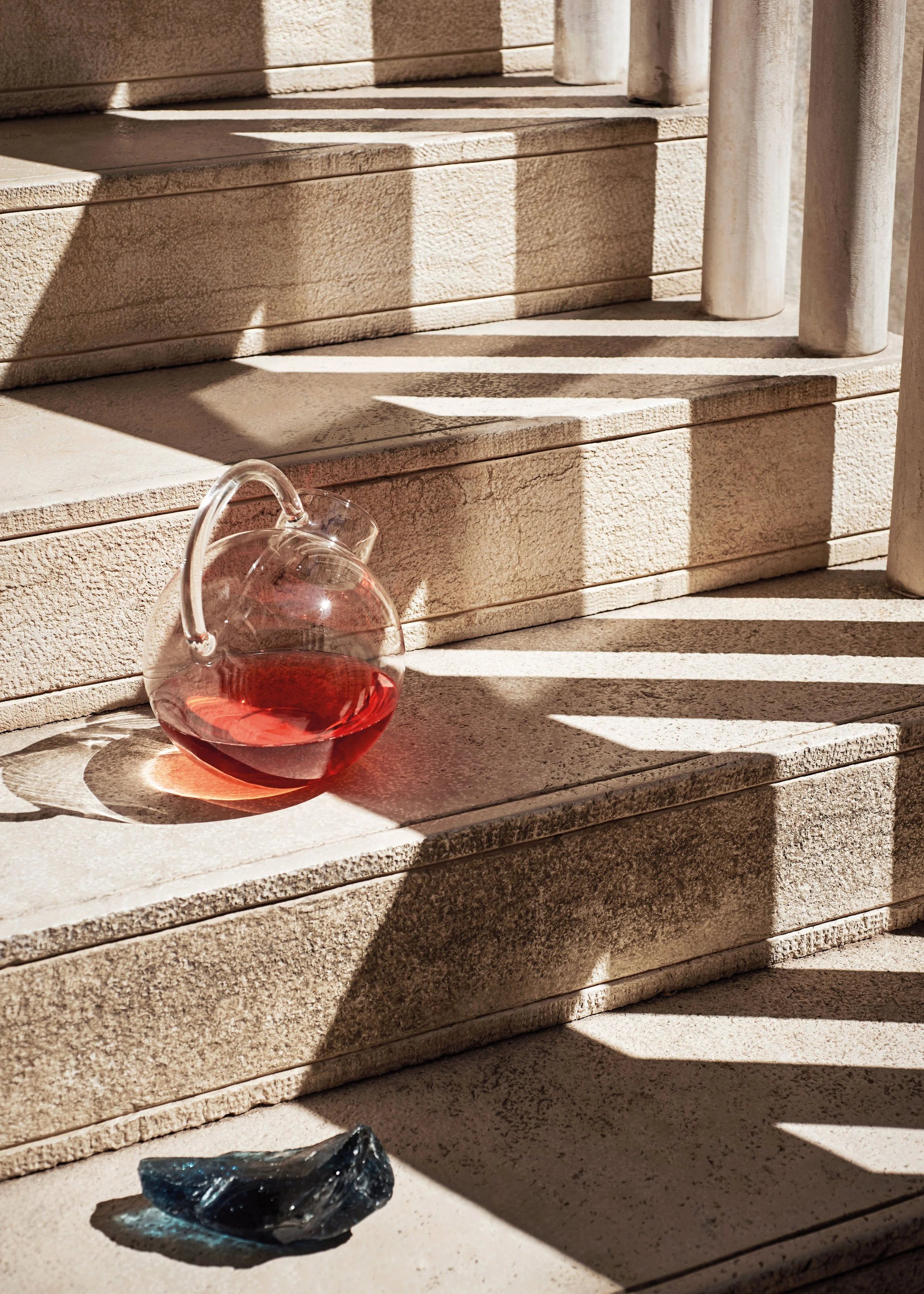Eine Glaskaraffe mit rotem Wasser steht auf einer Sonnenwärmten Treppe, daneben liegt ein dunkler Kristall.