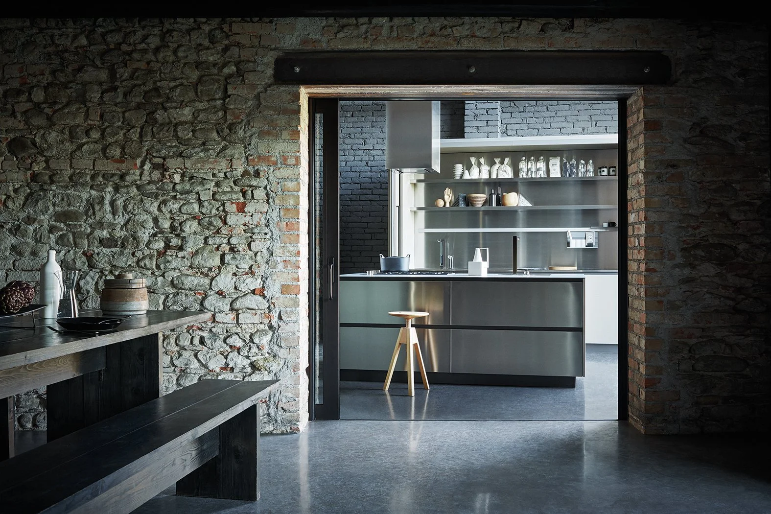 Moderne Küche im Industrial-Stil mit Edelstahl-Oberflächen, offener Regale und schwarzen Akzenten, sichtbar durch eine große offene Tür in einer Backsteinmauer