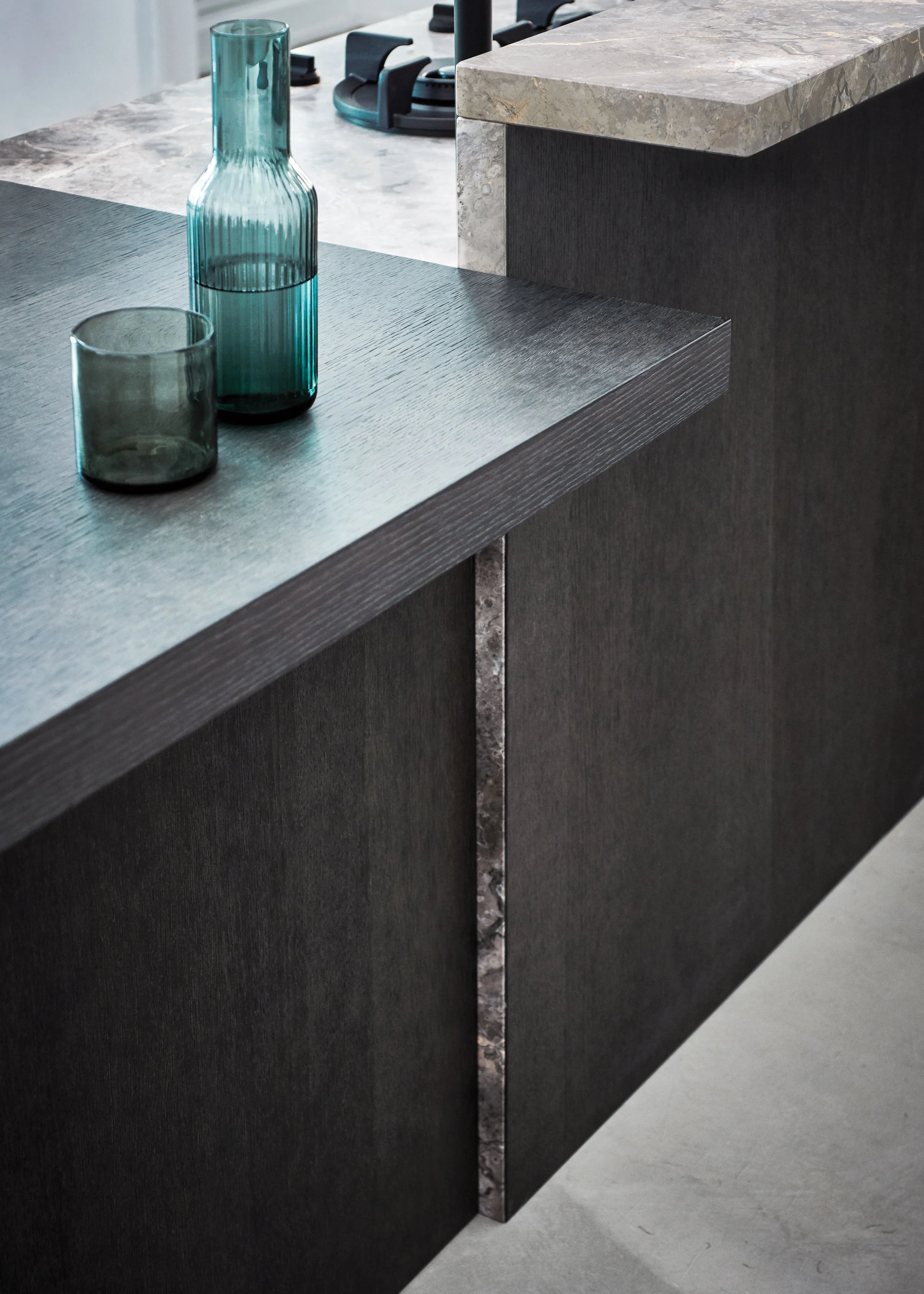 Nahaufnahme eines schwarzen Holzschranks mit dunkler Marmorplatte und metallischer Dekoration, daneben ein grauer Holztisch mit blauen Gläsern.