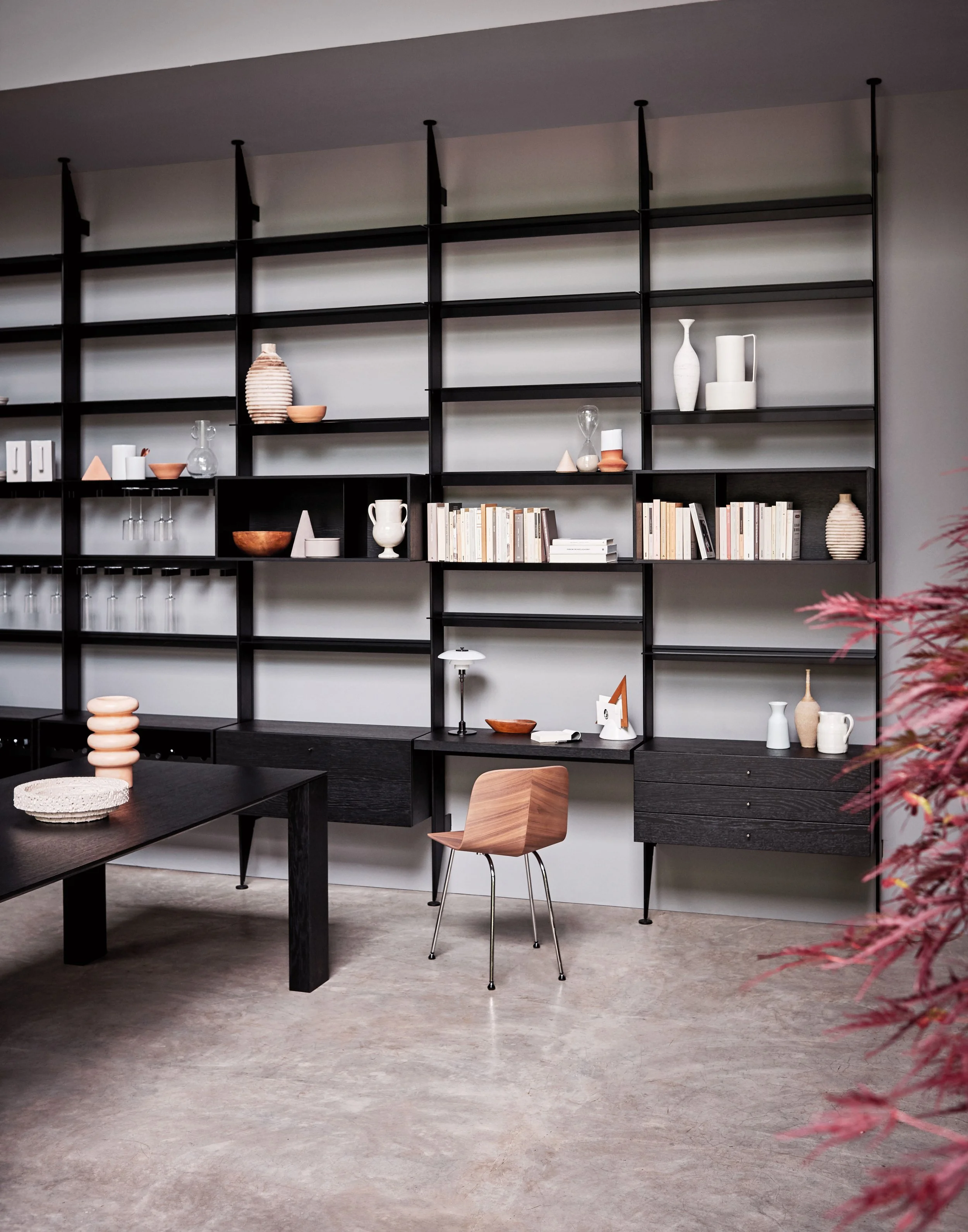 Schwarzes Bücherregal mit Dekorationsgegenständen wie Vasen, Kerzen, Büchern und Geschirr, im Hintergrund ein Esstisch und ein Holzstuhl, mit einer grauen Wand und einem grünen Teppich.