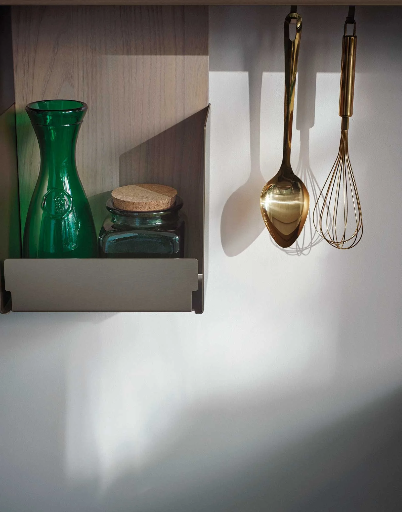 Bunte Glasvase, zwei Holzlöffel, ein Glas mit Korkdeckel sind an einer Wand im Schatten aufgehängt.