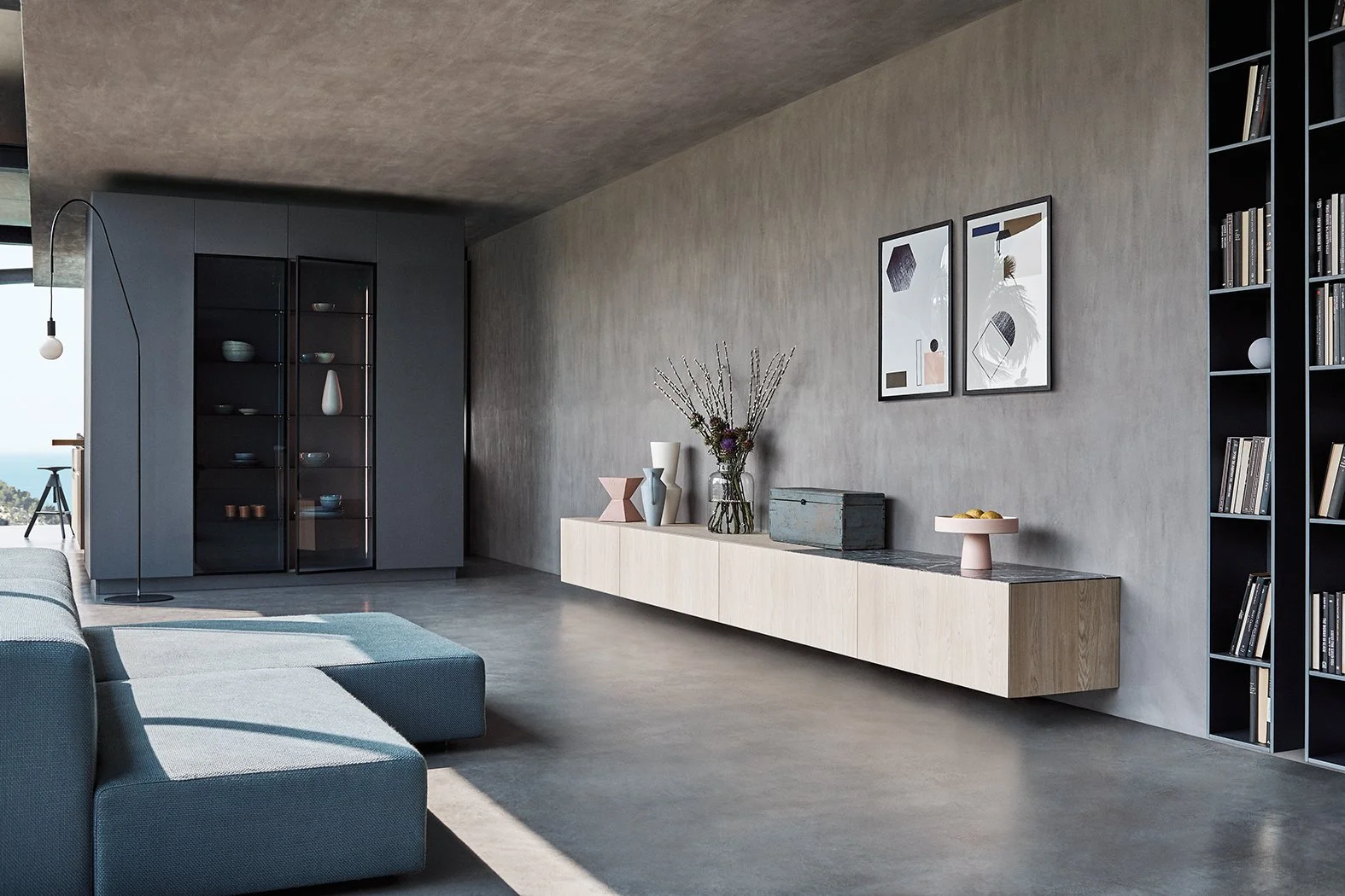 Moderner Wohnraum mit grauem Sofa, einer langen weißen Wandkonsole mit Dekoration, zwei modernen Bildern an der Wand, schwarzes Regal mit Büchern und einem Wohnzimmer mit großem Fenster, das Tageslicht hineinlässt.