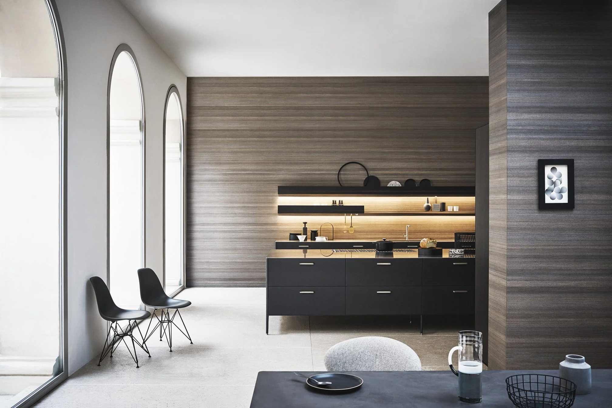 Moderne Küche mit eleganten schwarzen Küchenschränken, kombiniert mit einer natürlichen Holzwand und stilvollen Lampen als Beleuchtungselemente. Eine große Fensterwand lässt viel Tageslicht in den Raum und schafft eine helle, offene und luxuriöse Woh