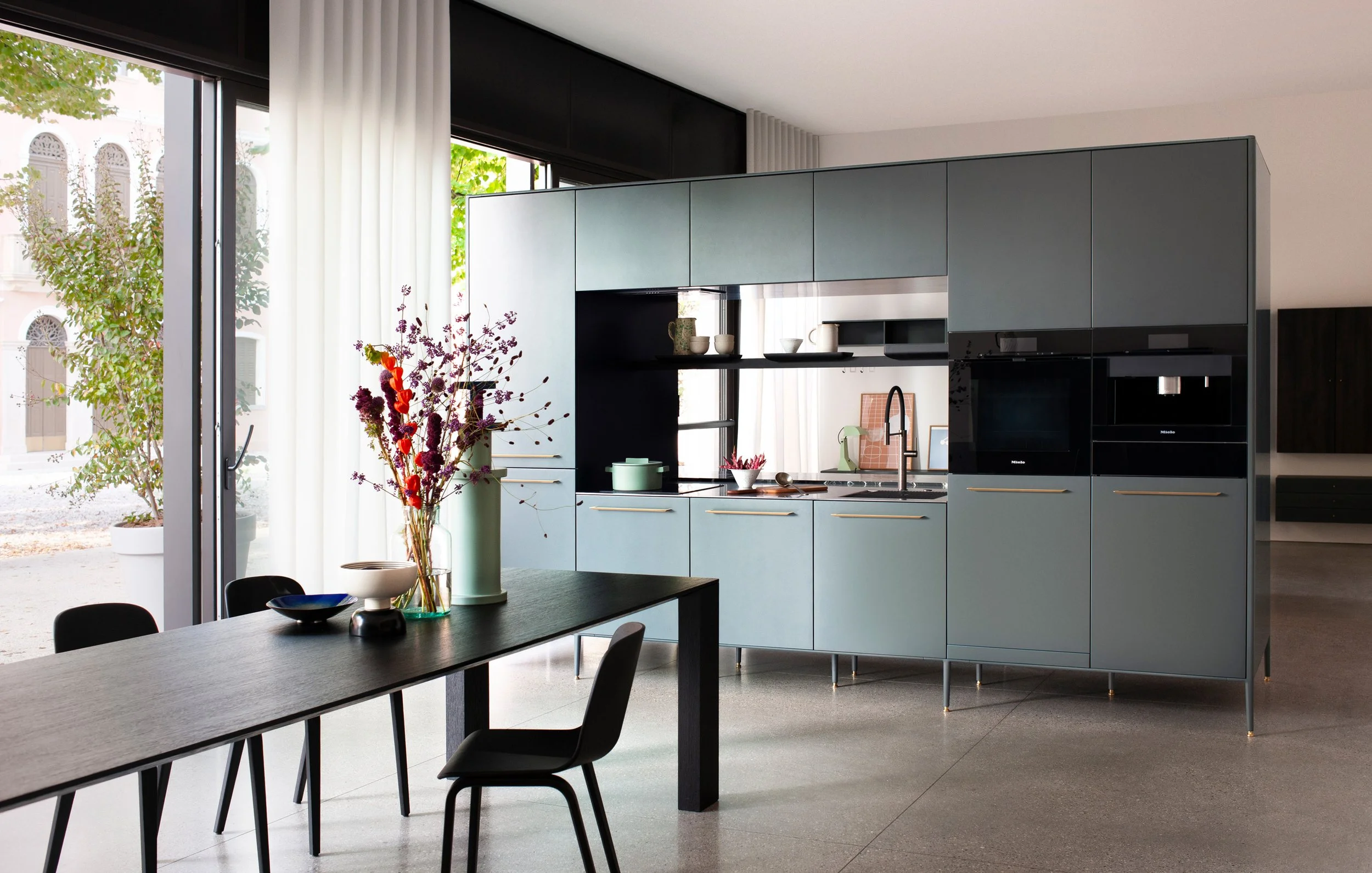 Moderne Küche mit schwarzen und blauen Schränken, offener Regale, einer Wasserflasche, bunten Blumen in einer Vase auf einem schwarzen Esstisch, große Fenster mit weißen Vorhängen, massiver dunkler Tisch und schwarze Stühle.