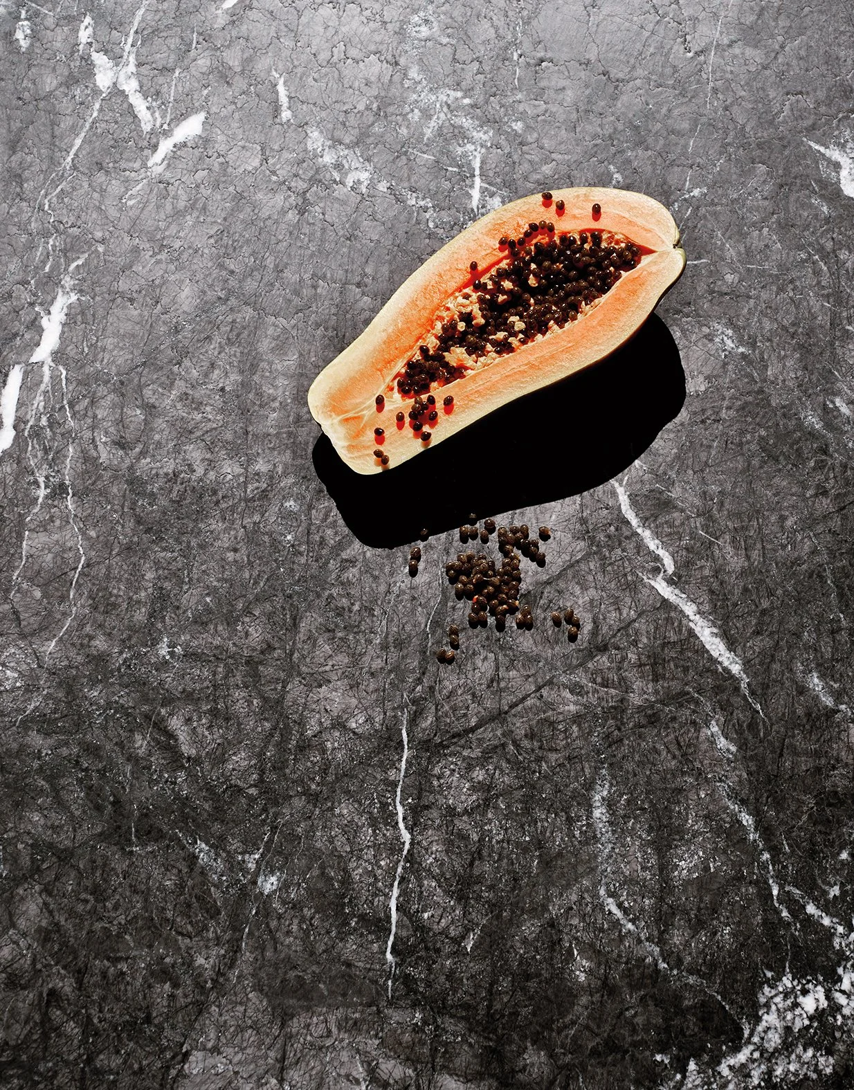 Eine halbe Papaya mit schwarzen Samen, einige Samen fallen auf eine dunkle Marmoroberfläche.