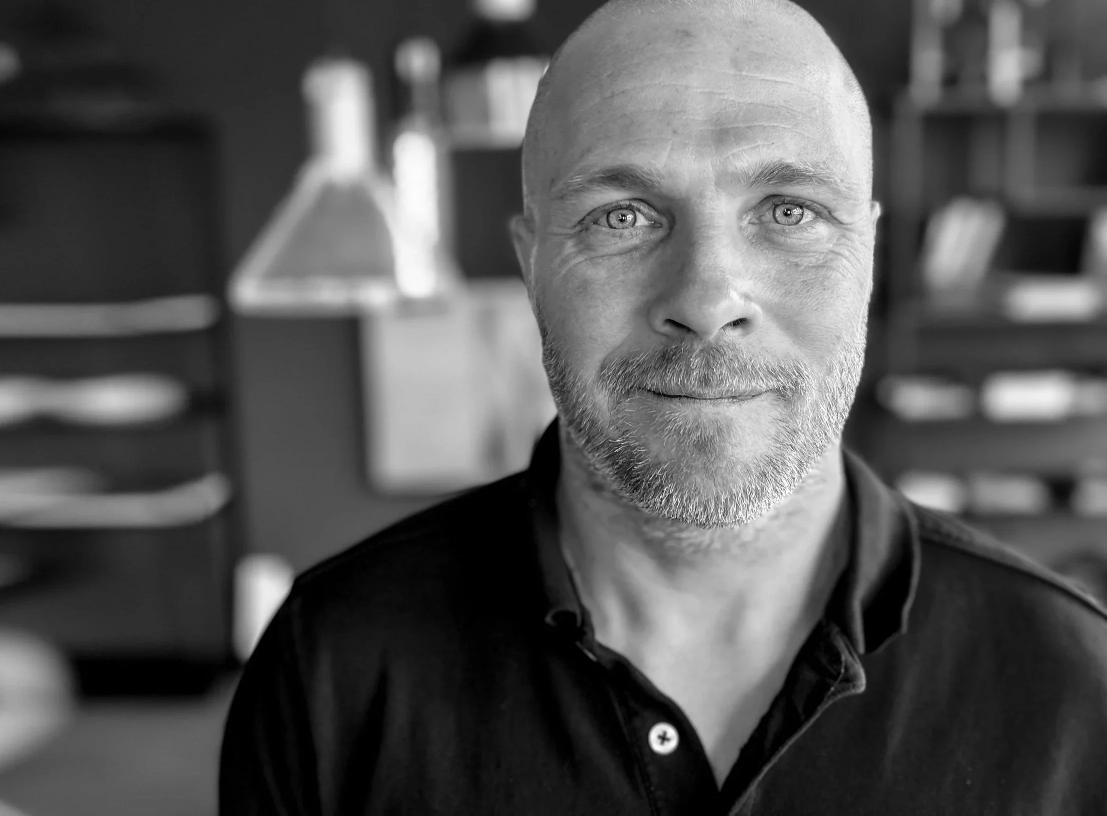 Schwarz-weiß-Foto eines Mannes mit runder Glatze und Bart, der in einem Labor mit Reagenzgläsern betrachtet. Im Vordergrund sind unscharfe Reagenzgläser.