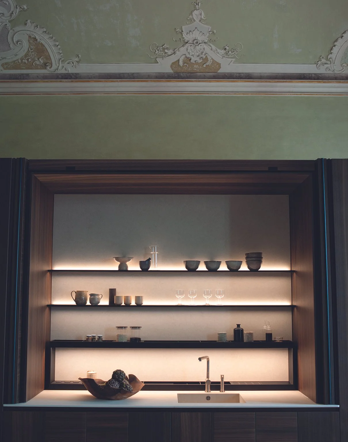 Moderne Küchenzeile mit offenem Regal, Keramik-Geschirr, Gläsern und Dekorationsobjekten, beleuchtet mit warmem Licht, unter einer traditionellen, kunstvoll verzierten Decke.