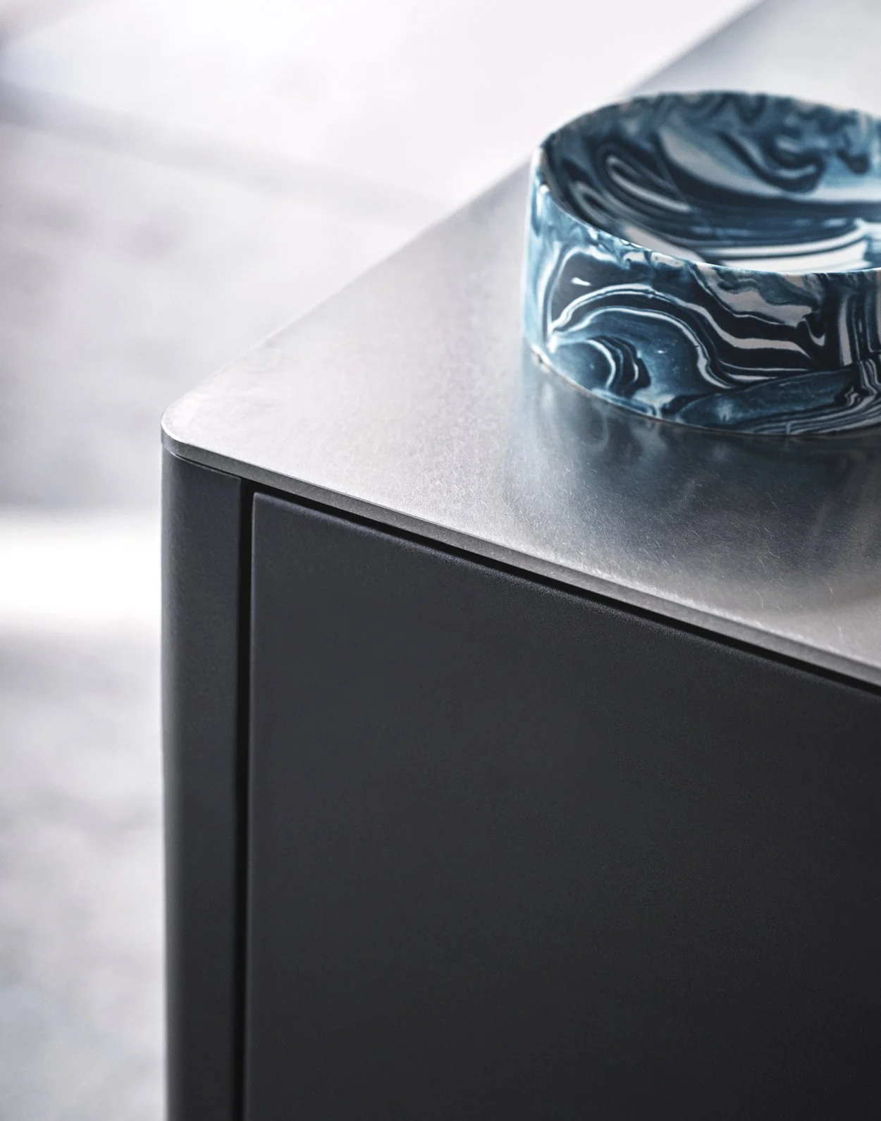 Ein eleganter blau-weiß marmorierter Kerzenhalter als modernes Designobjekt steht auf einer edlen silbernen Tischplatte und setzt einen stilvollen Kontrast zur minimalistischen schwarzen Möbelfront im Hintergrund. Die hochwertige und reduzierte Innen