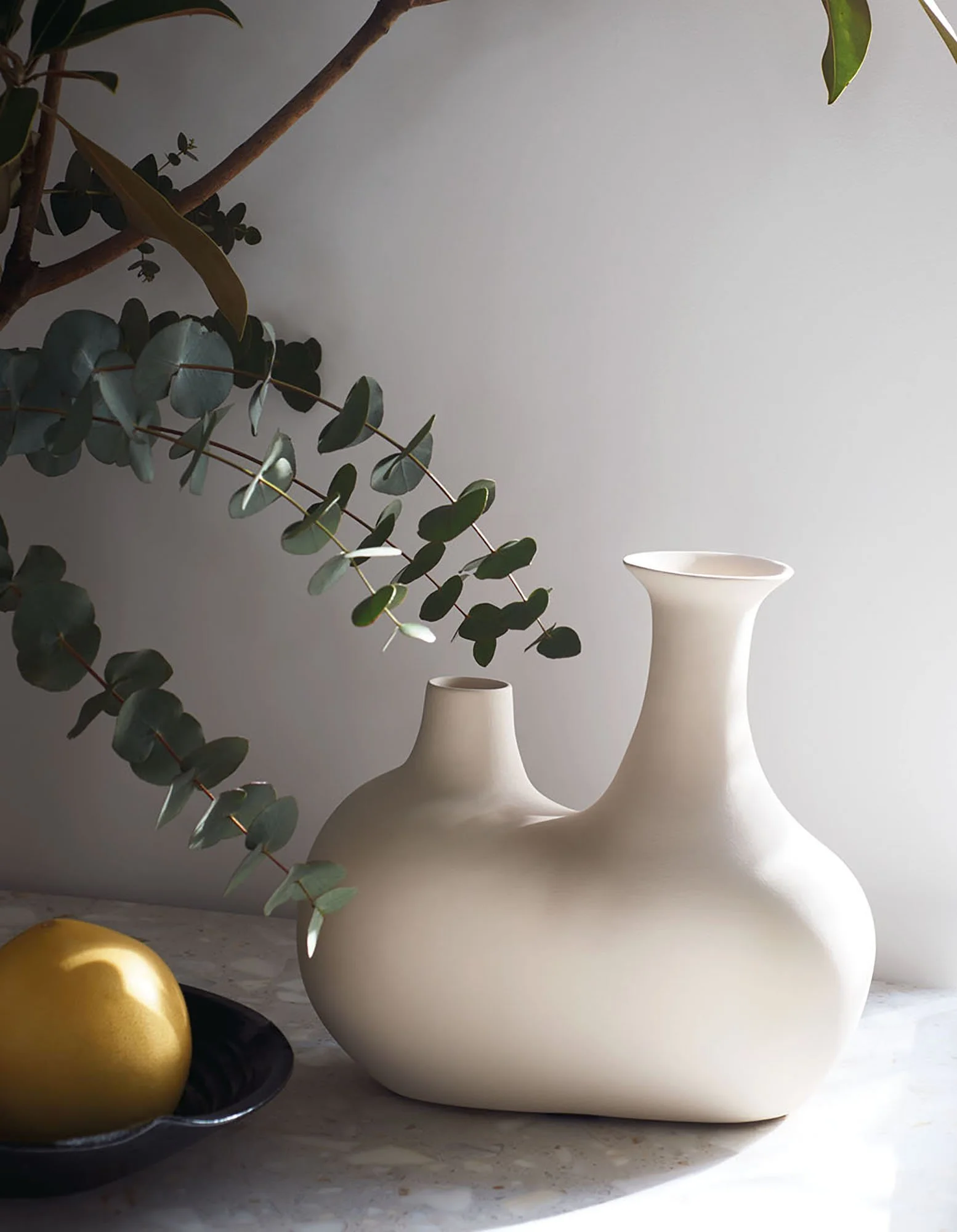 Weiße Keramikvase mit geschwungenem Hals, gefüllt mit grünen Blättern, auf einer hellen Oberfläche, daneben eine schwarze Schale mit goldenen Eiern, im Hintergrund ein grauer Hintergrund.