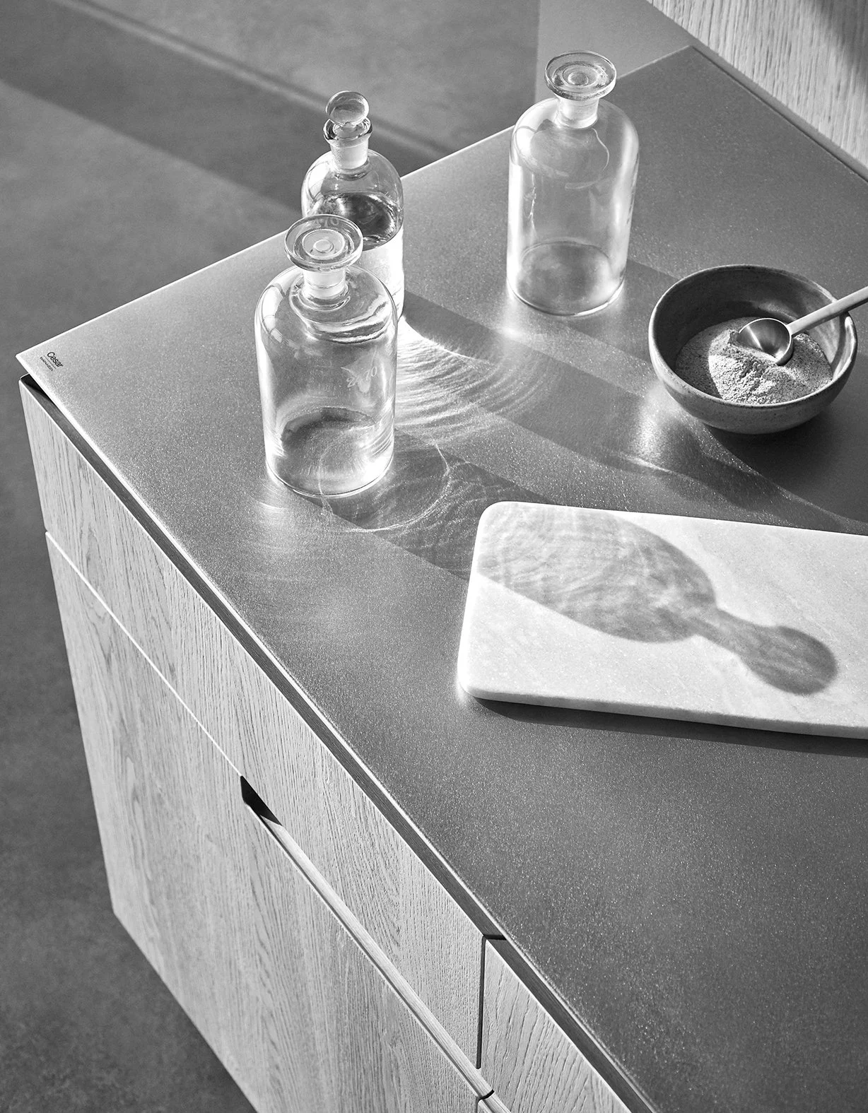 Schwarze und weiße Aufnahme eines modernen Holzschränkchens mit Gläsern, einer Schale mit Löffel und einem weißen Schneidebrett, auf dem ein Schatten eines Löffels mit einem Glas reflektiert wird.