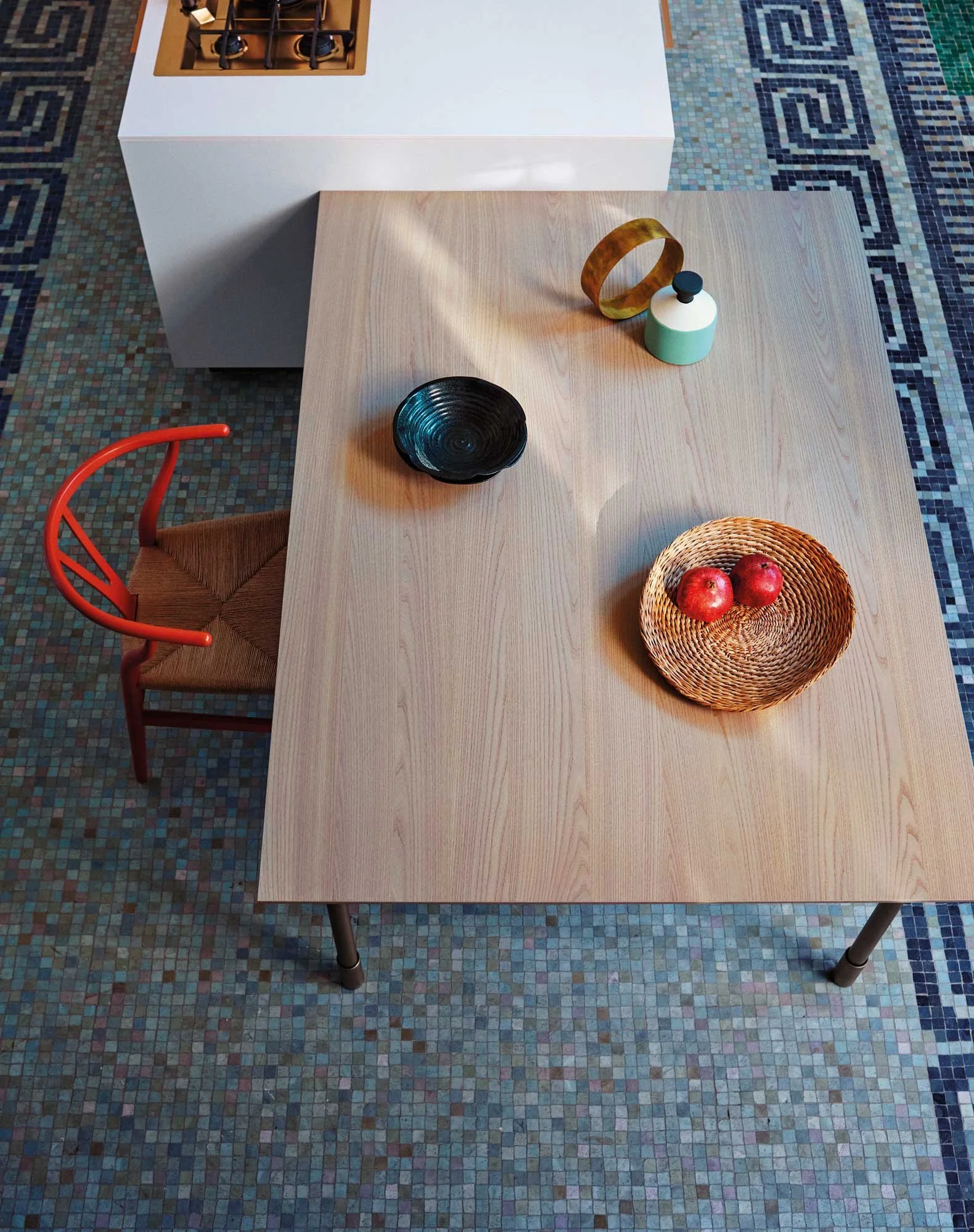 Ein Holztisch mit einer schwarzen Schale, einem Korb mit Äpfeln, zwei geometrischen Dekorationsgegenständen, und einem weißen Backofen in einem Raum mit buntem Mosaikboden.