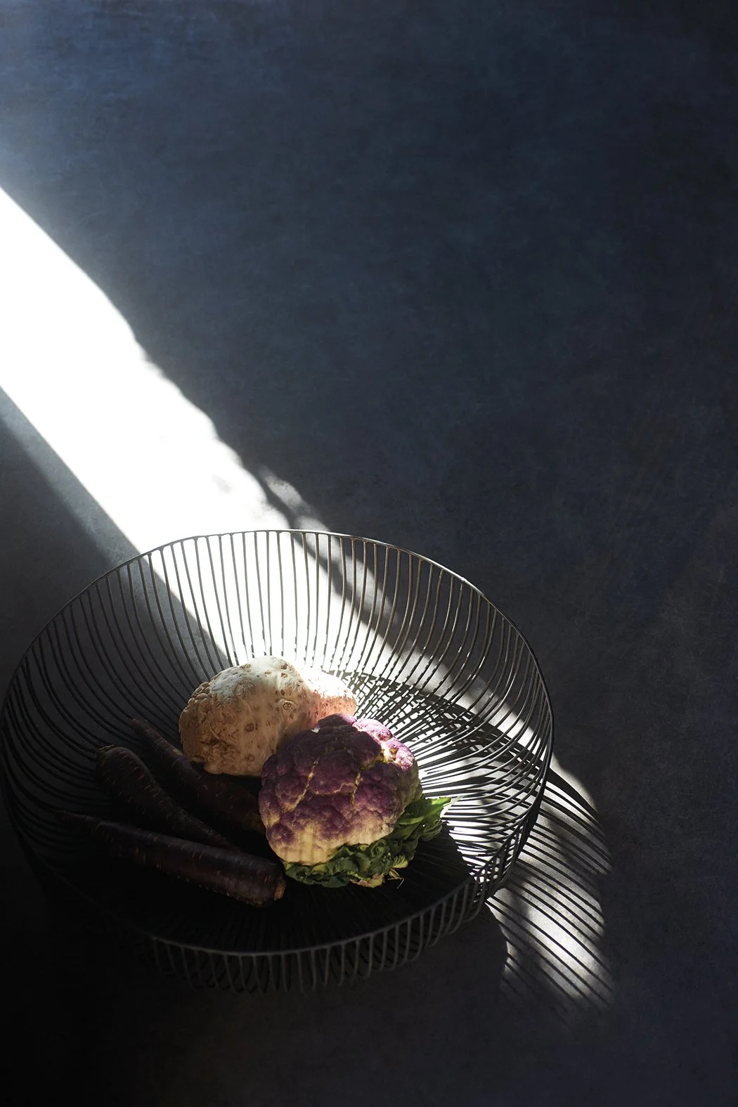Gemüse in einer runden Metalldrahtschale, darunter eine weiße, eine violette Blumenkohl und zwei lange, dunkle Möhren, im Gegenlicht fotografiert.
