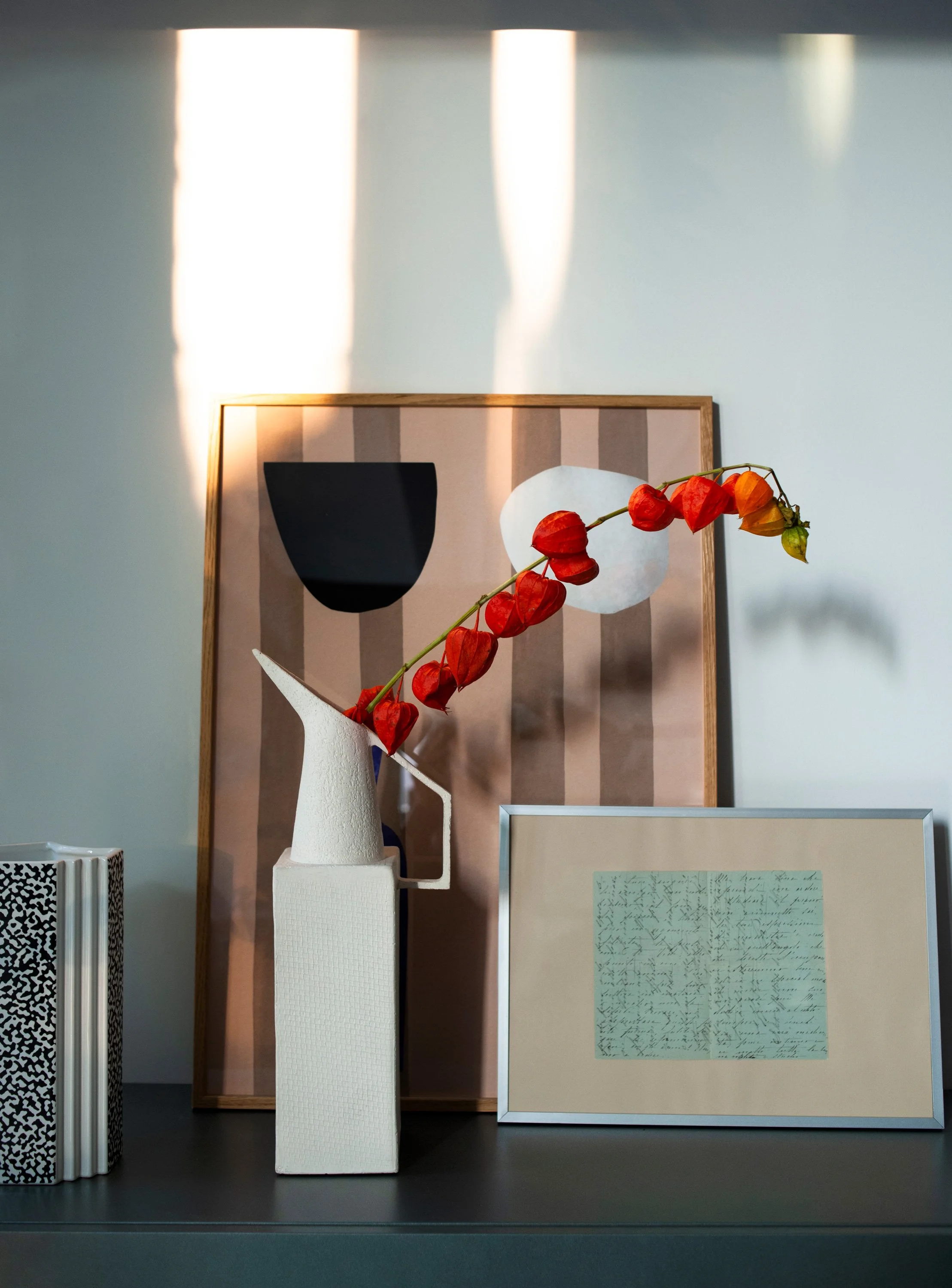 Ein stillleben mit einem weißen Keramik-Vase mit roten, thoraxförmigen Blumen, beleuchtet von Sonnenlicht, hinter einem gerahmten handgeschriebenen Brief auf grauem Tisch, umgeben von dekorativen Objekten auf einer dunklen Oberfläche.