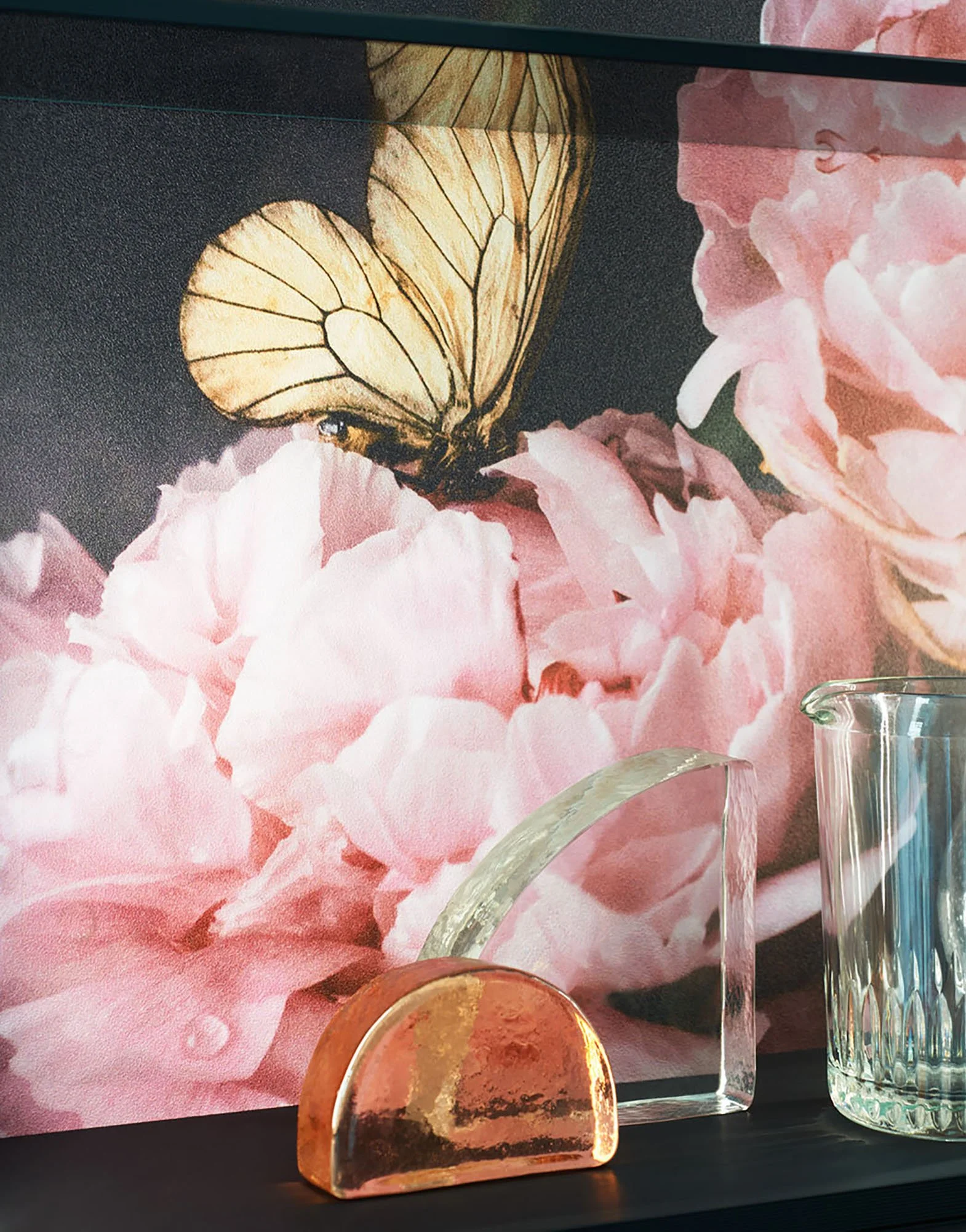 Eine Wand mit einem riesigen Wandbild eines gelben Schmetterlings auf rosa Blüten. Darunter stehen drei verschieden große Glasvasen auf einem schwarzen Regal.