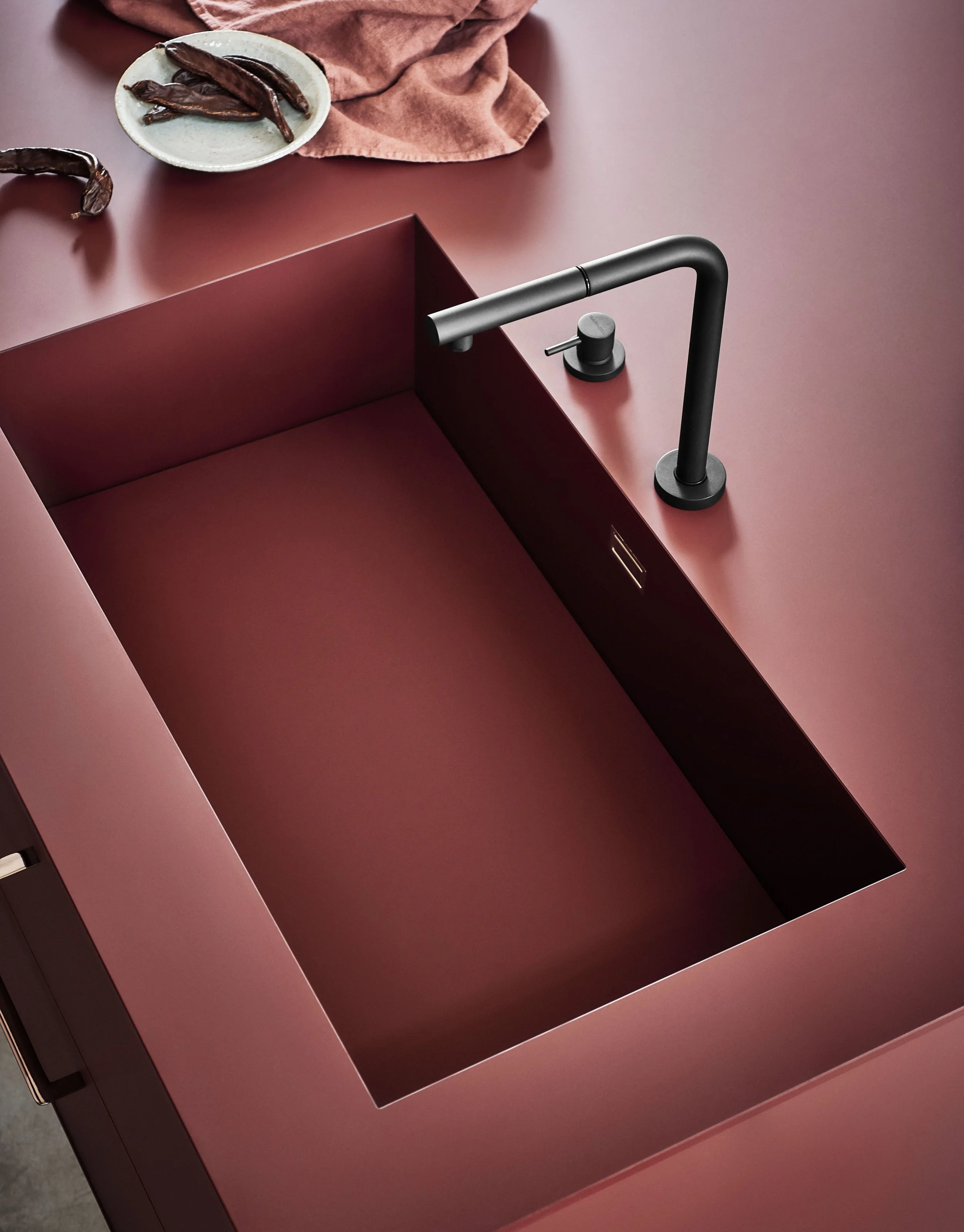 Ein roter Waschbecken mit schwarzer Armatur und einer weißen Schüssel mit getrockneten Chilischoten auf einem rosa Tuch.
