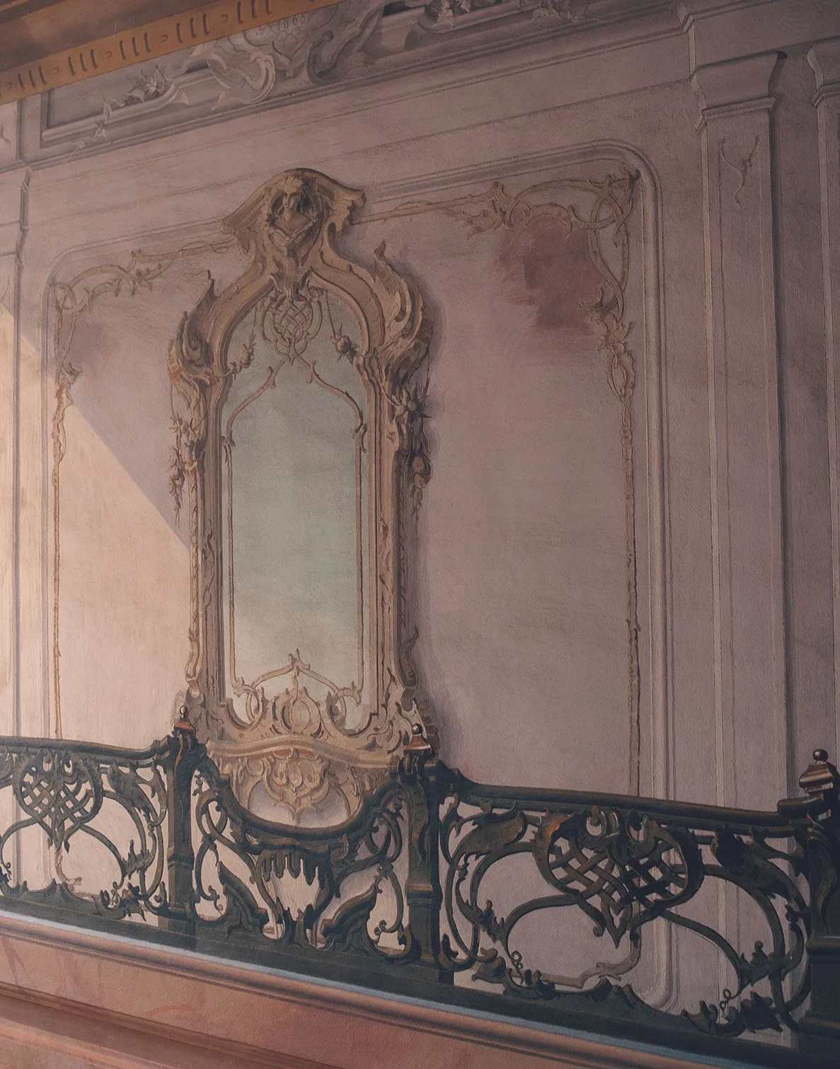 Ein aufwendig verziertes Wandbild mit einer großen, barockartig gestalteten Säule und einem Spiegel in der Mitte, ergänzt durch ein kunstvolles schmiedeeisernes Geländer im Vordergrund.