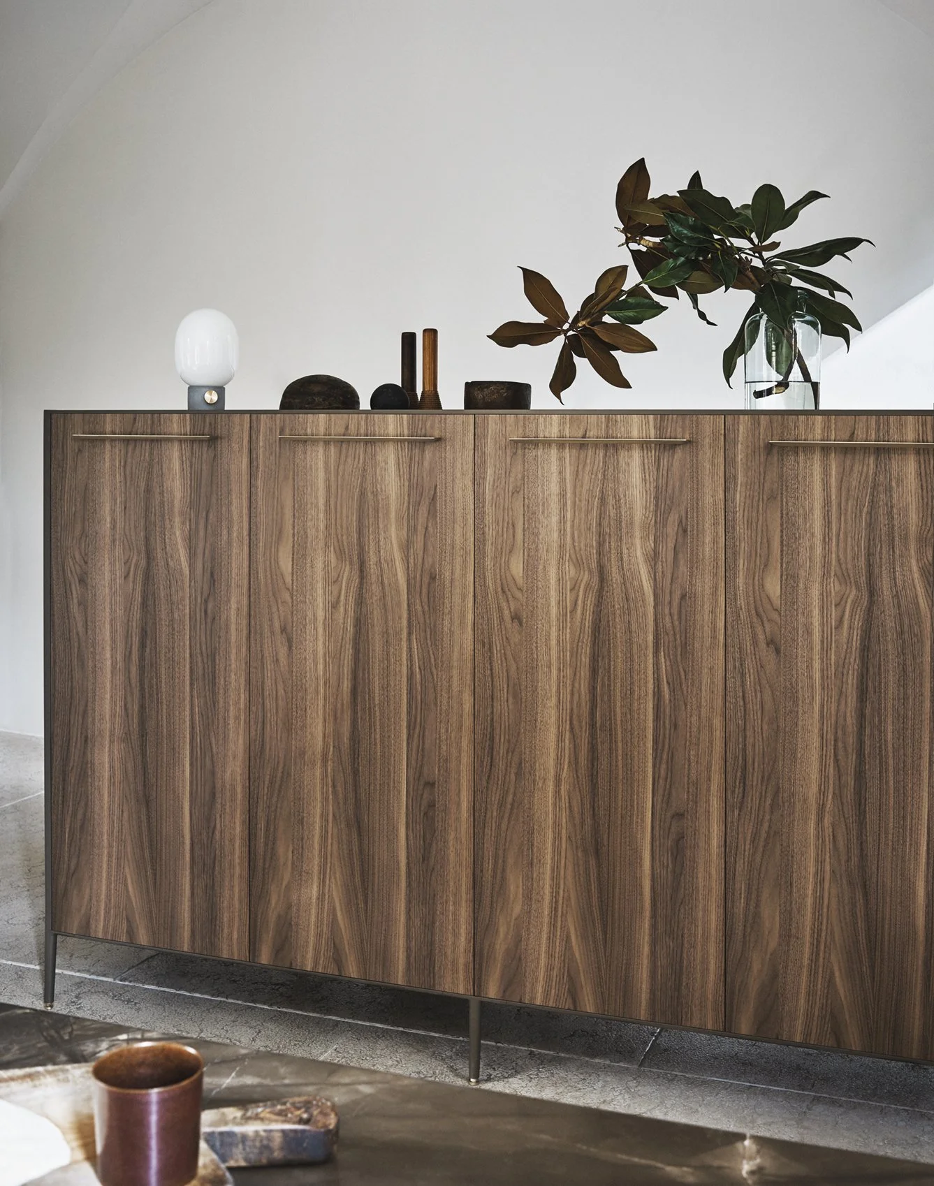 Eine Holzkommode mit schwarzen und braunen Dekorationsgegenständen sowie einer Glasvase mit großen grünen Blättern auf einer weißen Wand