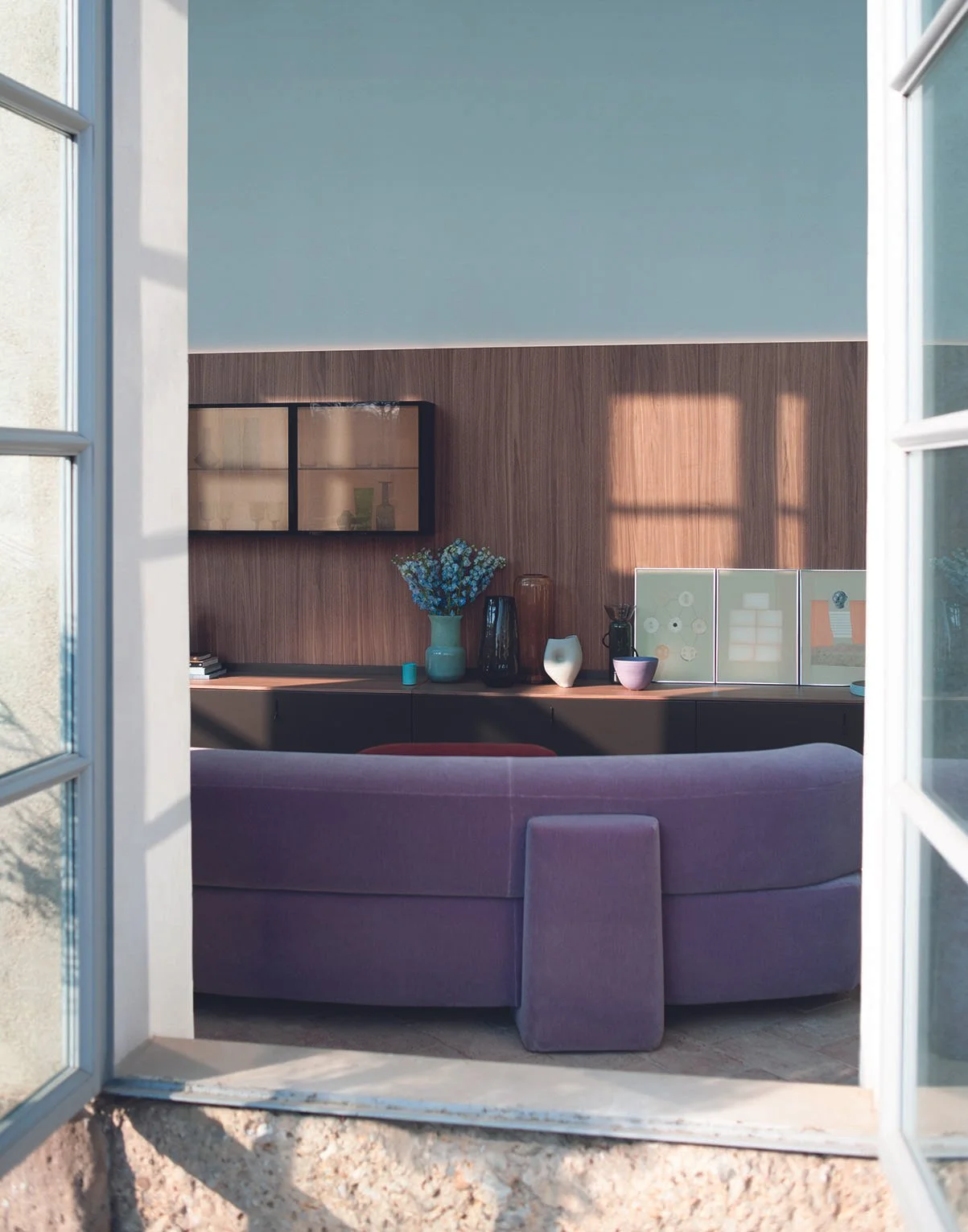 Der Blick in ein modern eingerichtetes Wohnzimmer durch ein offenes Fenster. Im Hintergrund eine Holzvertäfelung, auf der ein Wandregal mit Dekorationsgegenständen hängt. Auf einer Kommode stehen Vasen, Bücher, Dekorationsobjekte und Pflanzen. Im Vor