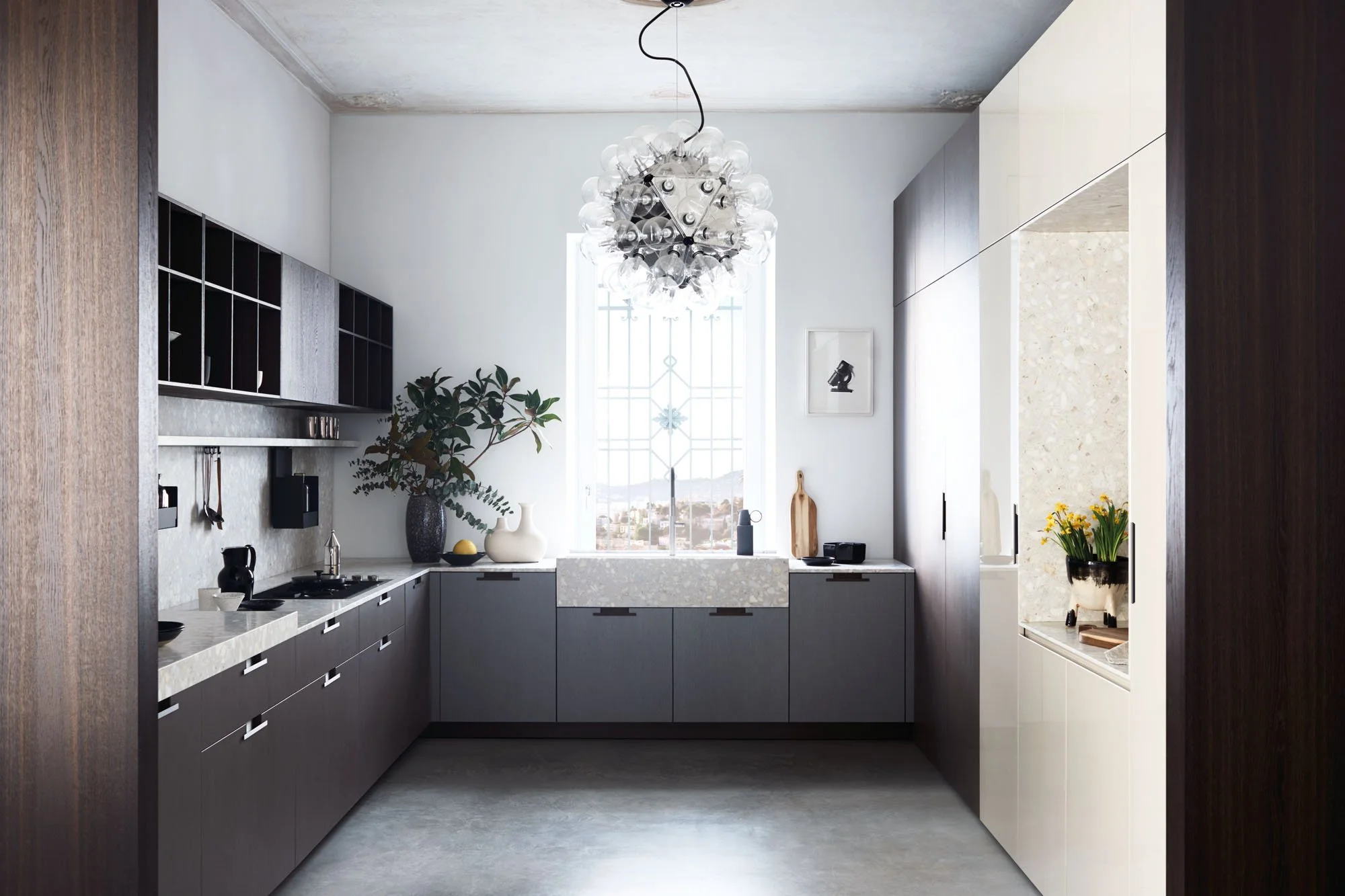 Moderne Küche mit weißen und grauen Schränken, einer großen Fensterfront, einer zentralen Küchenspüle aus Marmor, Küchenutensilien und Dekorationsgegenständen, einem runden Kronleuchter und einigen Pflanzen.