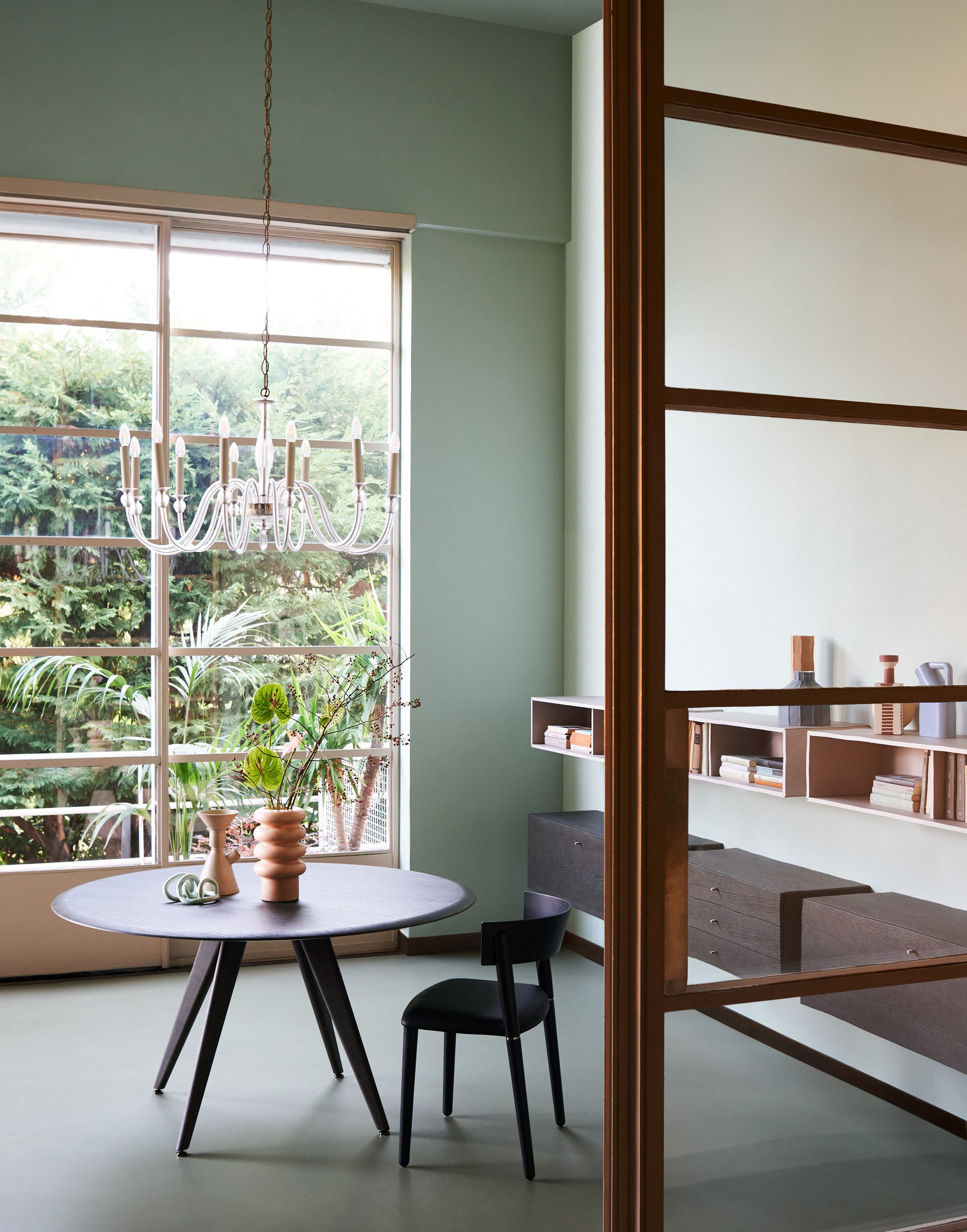 Ein elegantes Esszimmer mit einem runden schwarzen Tisch, zwei schwarzen Stühlen, einer dekorativen Vase, einem Wandregal mit Büchern, großen Fenstern mit Blick nach draußen auf grüne Bänke und Pflanzen, sowie eine weiße Kronleuchterlampe. Der Raum h