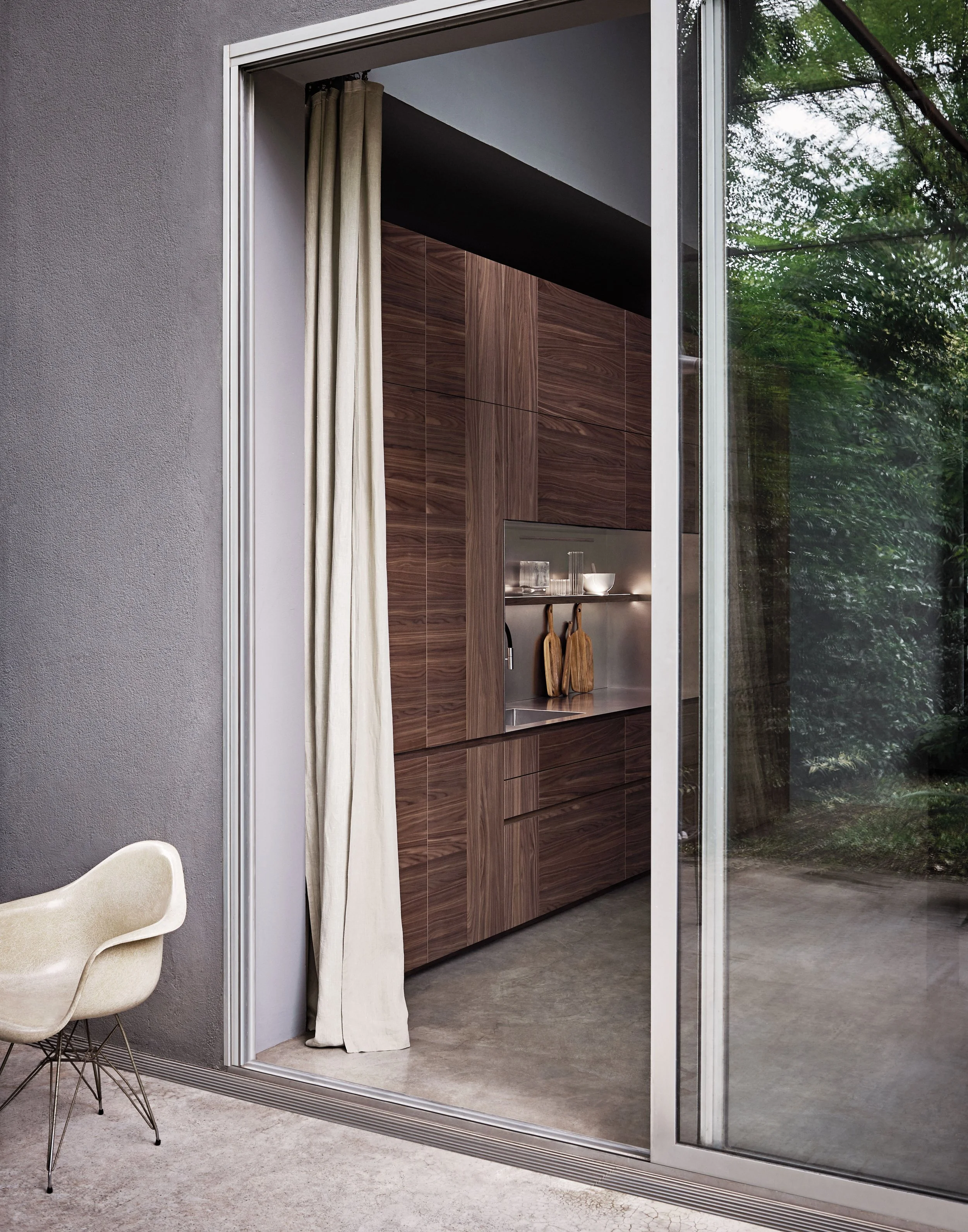 Innenansicht eines modernen Wohnzimmers mit großem Fenstertüren, weißen Stühlen und einer Küche mit Holzfronten im Hintergrund, Blick nach draußen auf grüne Bäume.