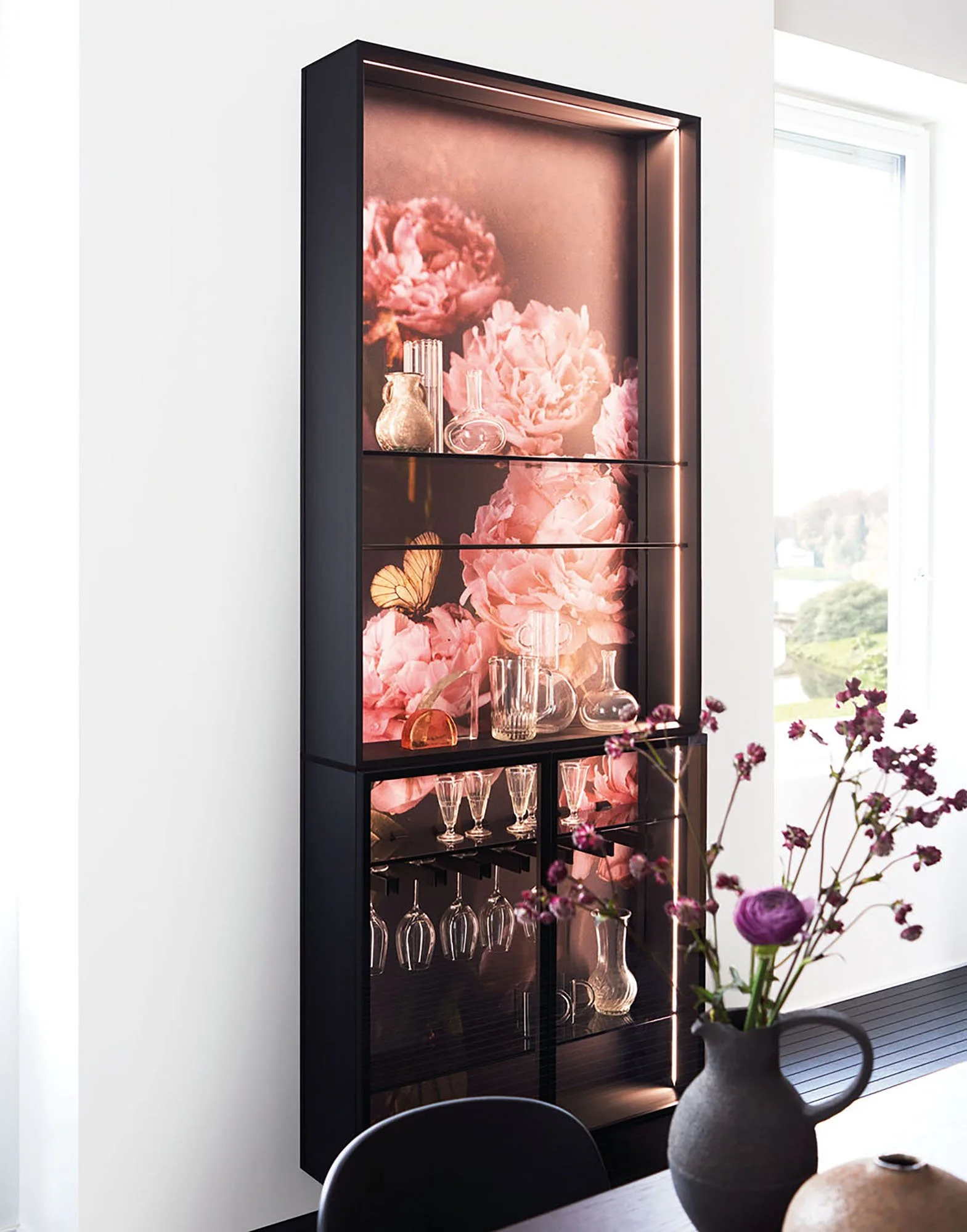 Ein schwarzes Sideboard mit Glastüren, das mit Dekorationsglaswaren und pinken Blumen hinter beleuchtetem Glas dekoriert ist, neben einem Fenster mit Blick auf eine grüne Landschaft und einer Vase mit violetten und weißen Blumen.