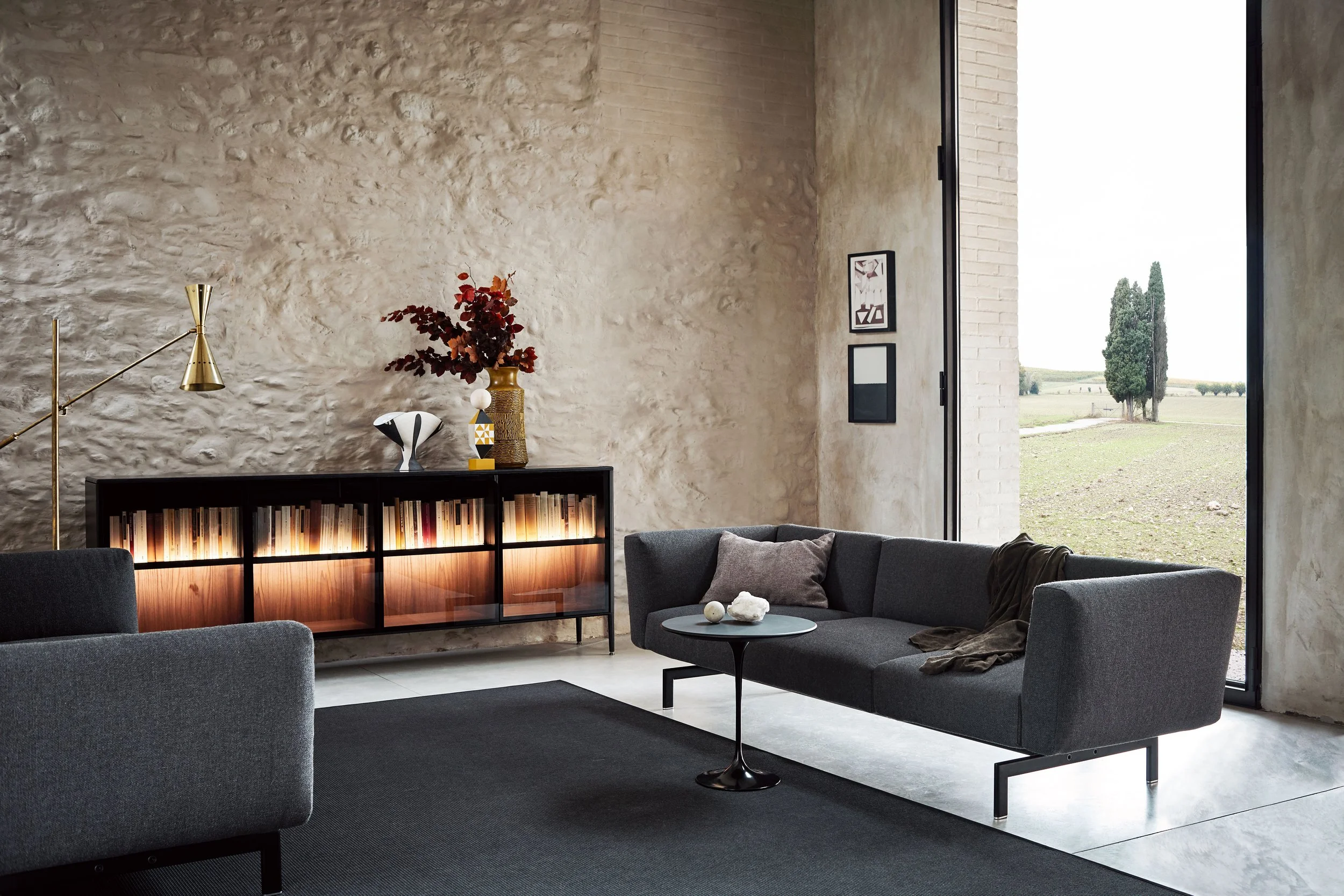 Moderner Wohnraum mit großen Fenstern, grauen Sesseln, einem schwarzen Sideboard mit Büchern, Dekorationsobjekten und Bildern, sowie einer stilvollen Stehlampe und Tischdeko.