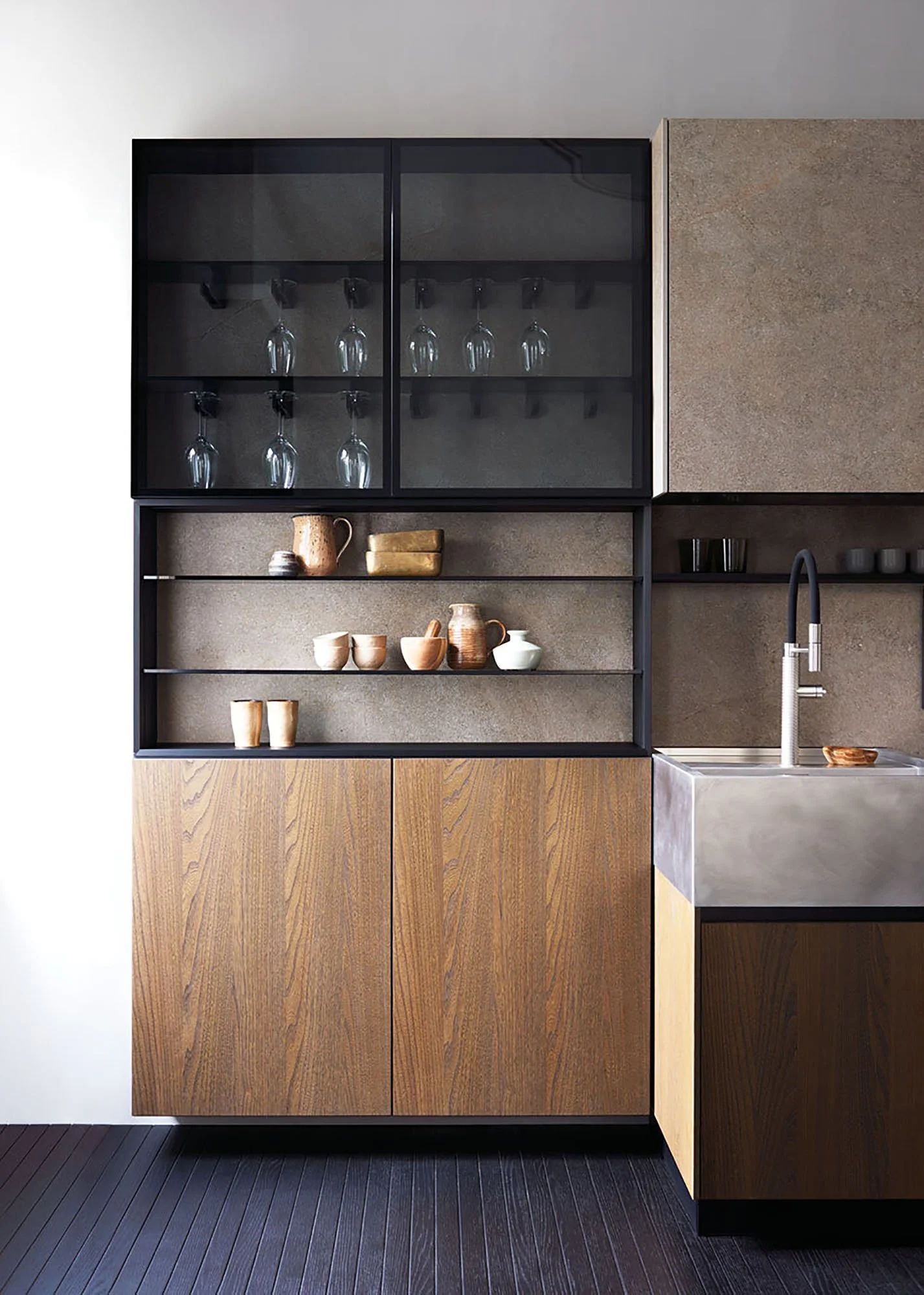 Moderne Küche mit schwarzen Regalen, Holzkasten und einem Edelstahlspülbecken. Es gibt offene Regale mit Keramik- und Holzgeschirr sowie Wein- oder Likörgläser in einem schwarzen Regal. Die Küche hat einen grauen Backstein-Wandhintergrund und dunklen