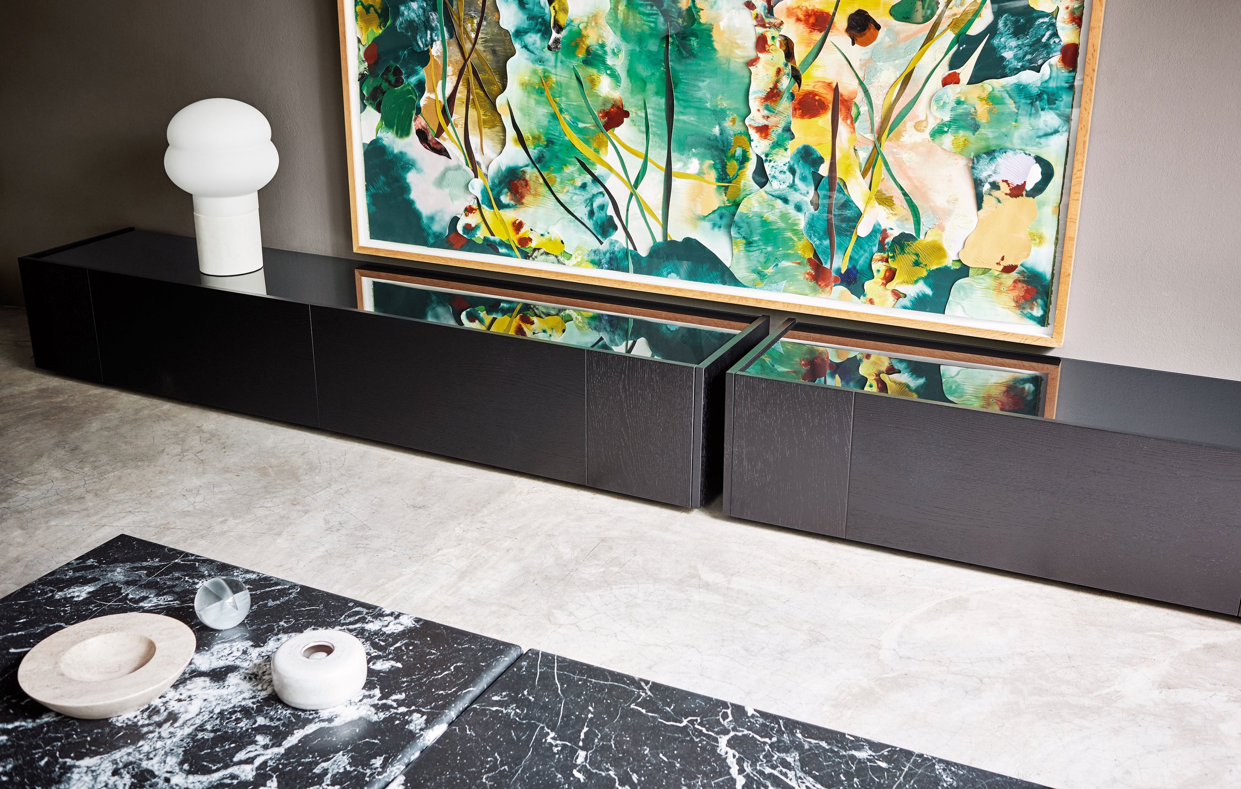 Ein modernes Wohnzimmer mit schwarzem Lowboard, einer großen abstrakten Wandmalerei mit grünen, gelben und rot-orangen Farben, einer weißen skulpturalen Vase auf dem Lowboard und einem schwarzen Tisch mit schwarzen und weißen dekorativen Objekten auf