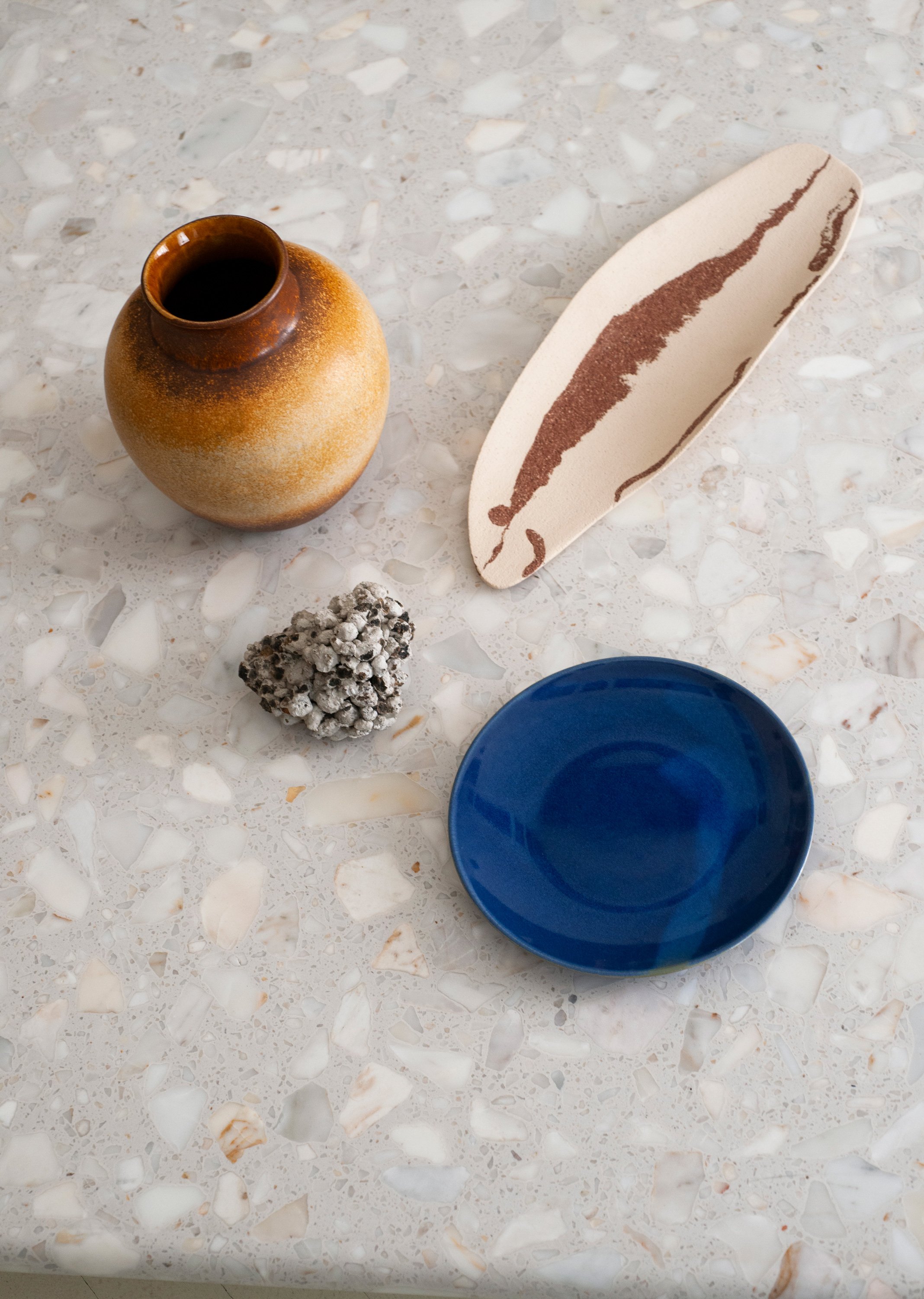 Tisch mit einer braunen Vase, einer weißen Platte mit braunem Muster, einem blauen Schüssel und einem kleinen Gebilde aus einer Steinformation.