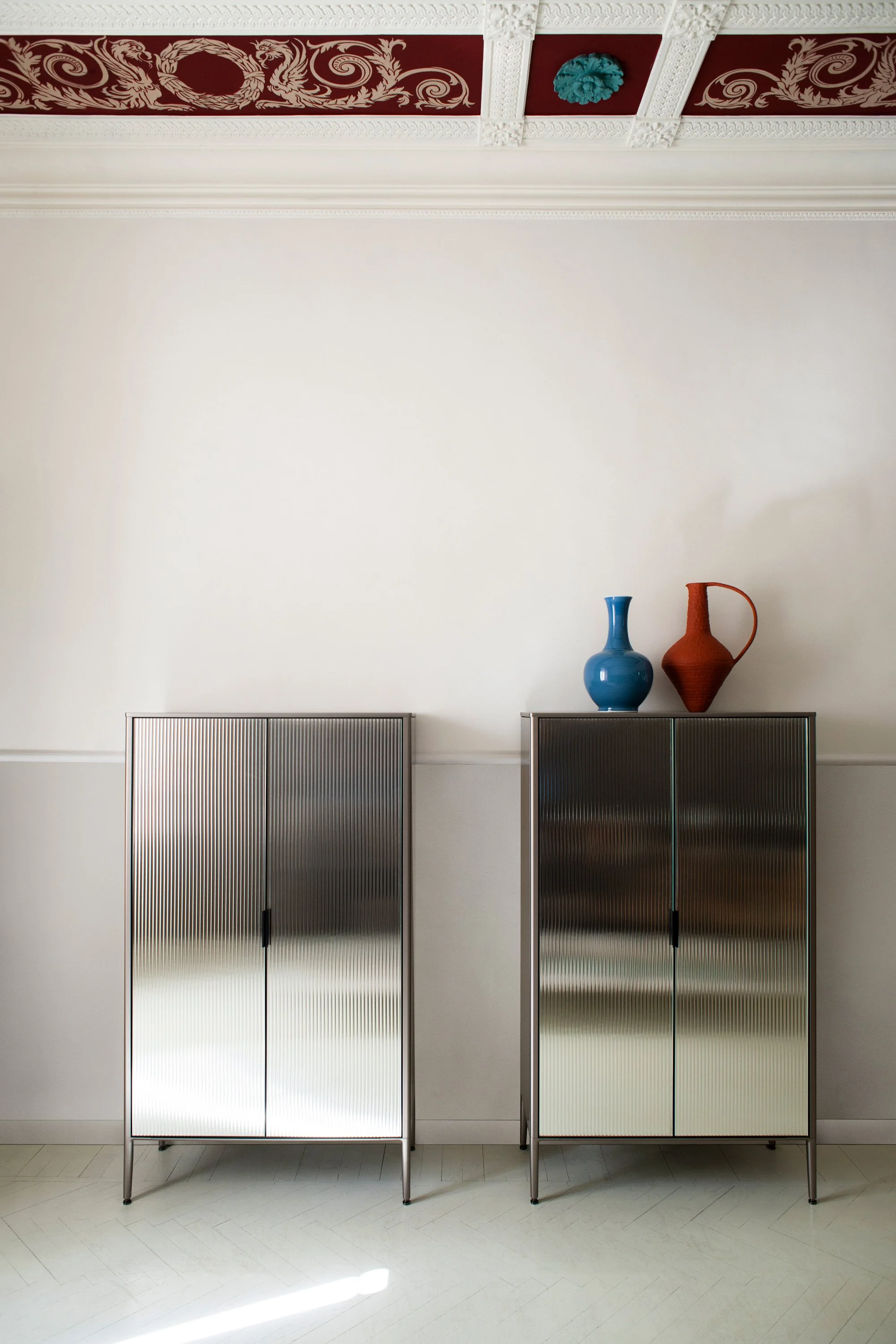 Zwei metallene Schränke mit vertikalen Linien, oben stehen zwei Vasen, eine blau und eine rot, vor einer weißen Wand mit dekorativen Zierleisten und einem Dekor im oberen Bereich.