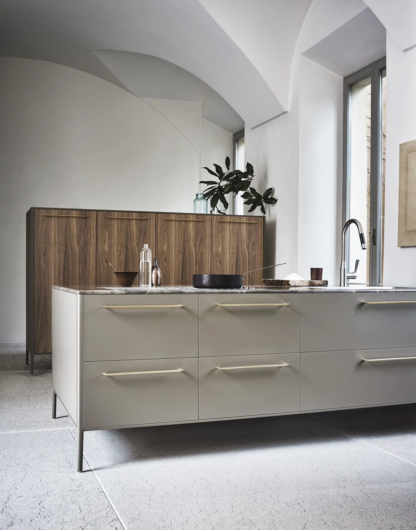 Moderne Küche mit weißer Insel, Holzschrank im Hintergrund, große Fenster, Wasserhahn und einige Dekorationen auf der Insel.