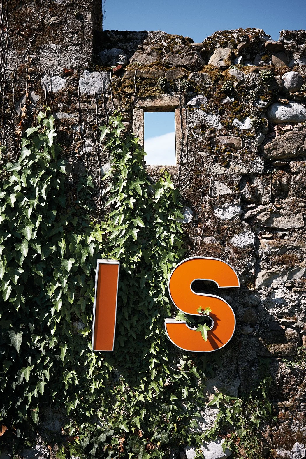 Ein altes Mauerwerk mit einem kleinen rechteckigen Fenster und grünem Efeu, das an der Mauer wächst. Darunter hängen große, orangefarbene Buchstaben, die das Wort 'IS' bilden.